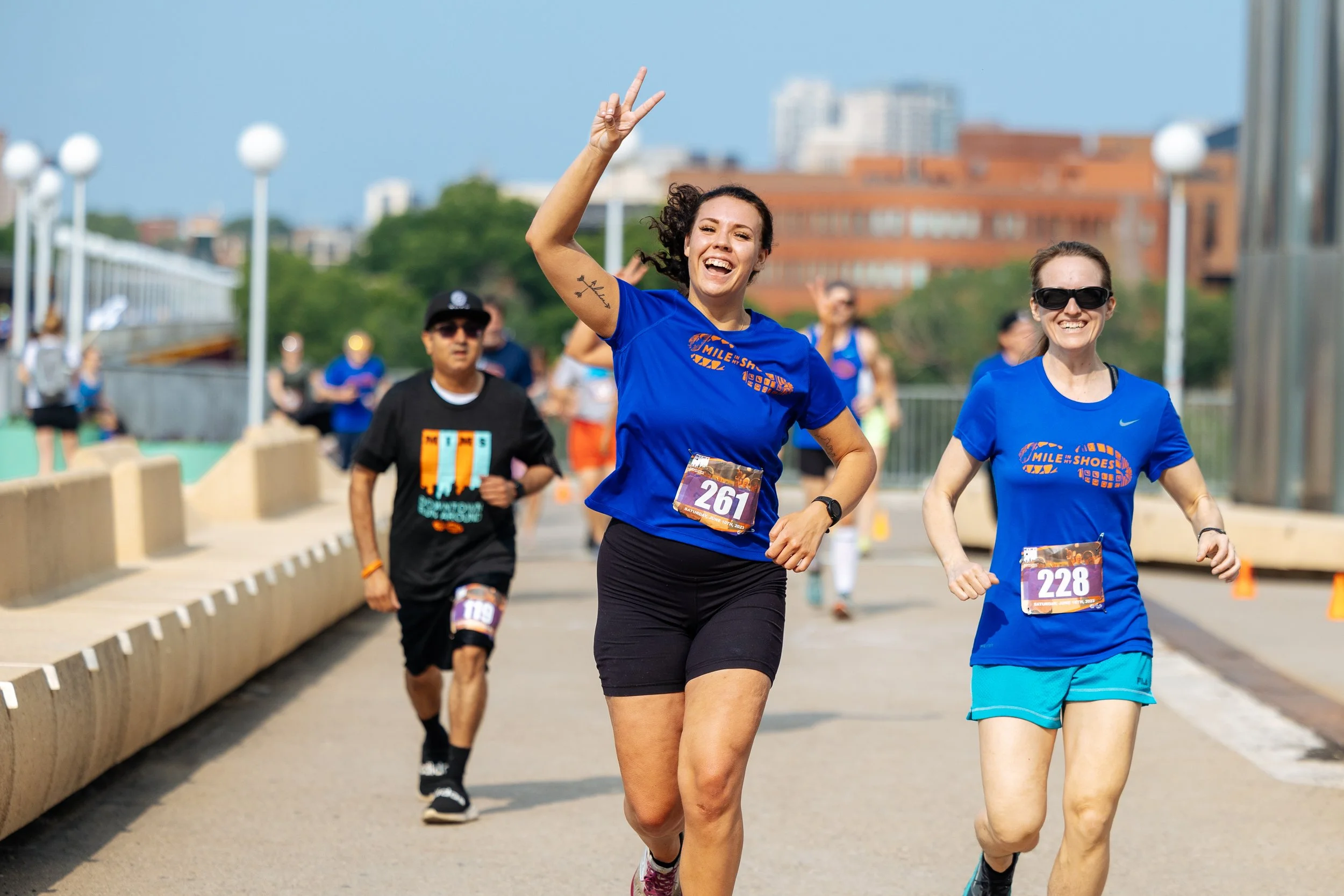 Group of runners participating in a marathon or race, smiling and enjoying the event on a sunny day, with urban buildings in the background.