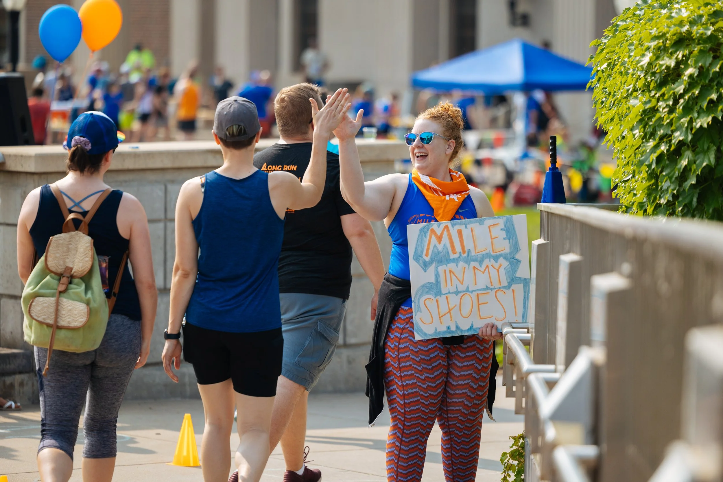 A woman wearing sunglasses and orange bandana high-fives a man at a community event. The woman holds a sign that says 'MILE IN MY SHOES.' There are other people and balloons in the background, indicating a festive outdoor gathering.
