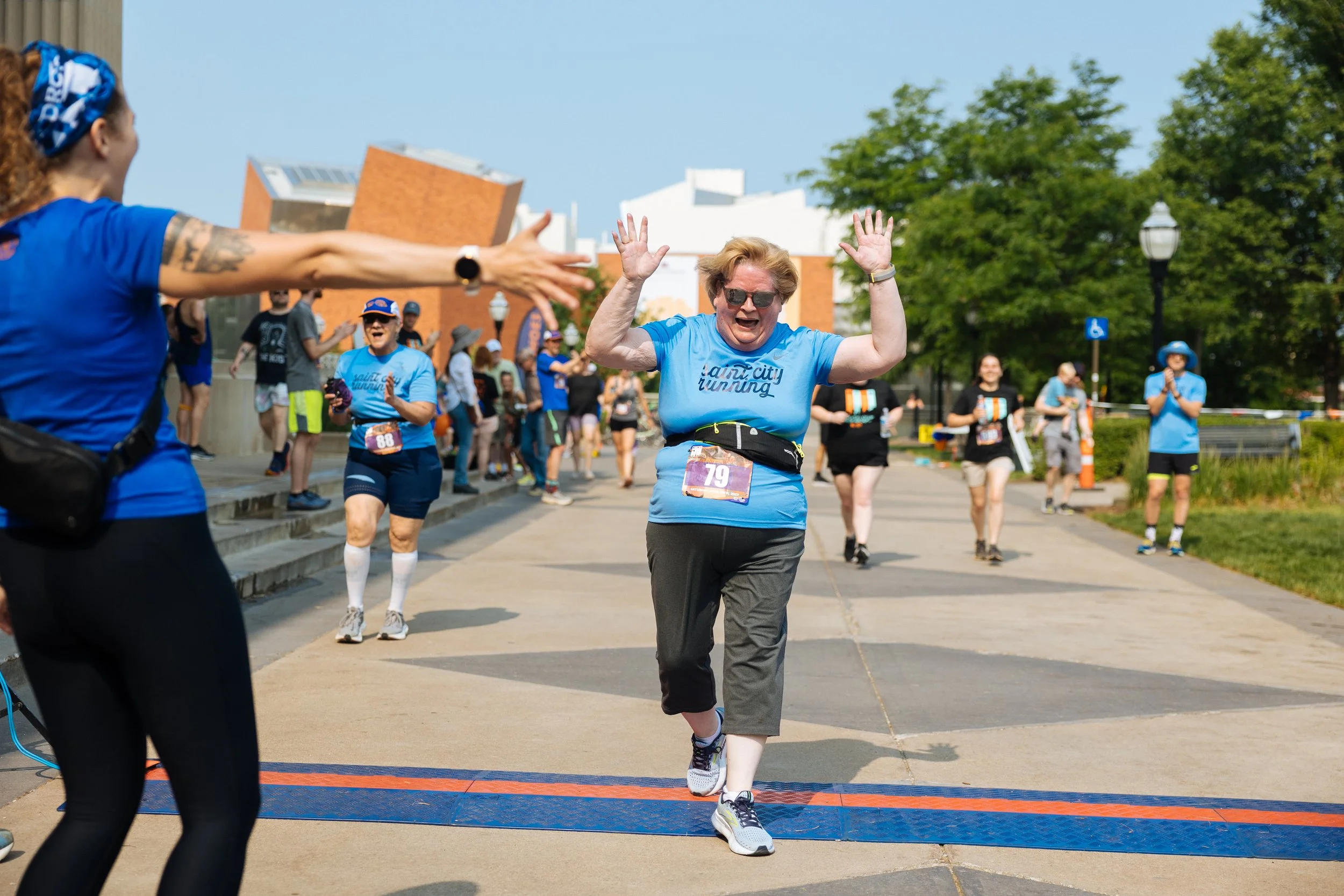 A woman in a blue shirt and gray pants crossing the finish line in a race, raising her hands in celebration, with other runners and volunteers in the background on a sunny day.