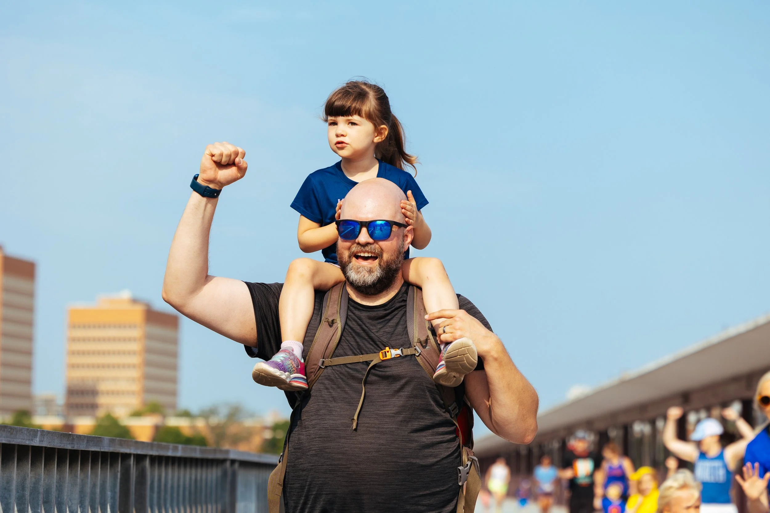 A man with a beard wearing sunglasses, a black shirt, and a backpack is carrying a young girl on his shoulders. The girl has brown hair, is wearing a blue shirt, and looks to the side with her hand resting on the man's head. They are outdoors on a su
