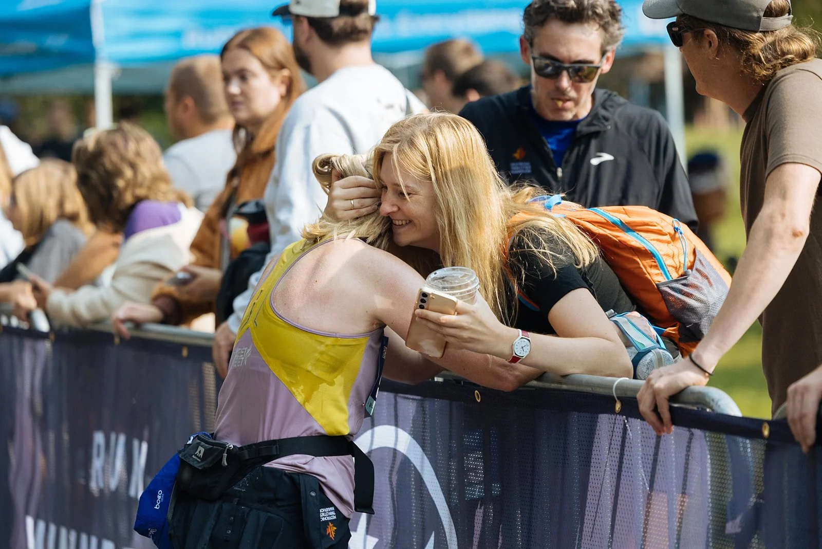 A woman with blonde hair, wearing a yellow and purple running tank top, hugs a smiling woman with long blonde hair, black shirt, and an orange backpack, as they lean on a barrier at a race event. The woman in the race is holding a phone and a plastic