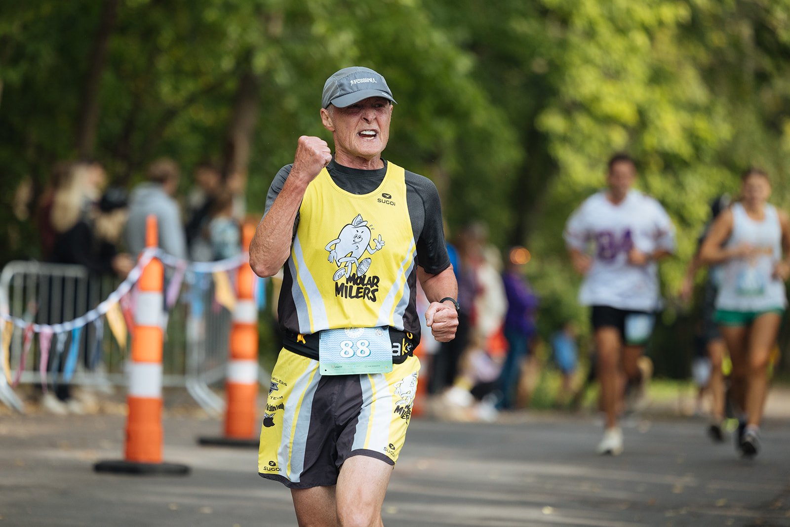 An older man running in a marathon, wearing a yellow and black sleeveless shirt with a cartoon character and the words 'Mullard Milers,' and a gray cap, surrounded by other runners and spectators in a park.