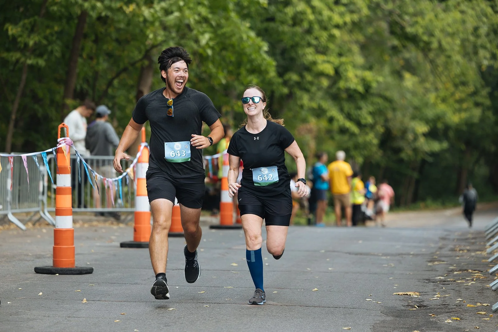 Two runners, a man and a woman, participating in a race on a paved road surrounded by trees. The man is wearing a black shirt, black shorts, and has sunglasses hanging from his shirt, with a race number 643. The woman is wearing a black shirt, black 