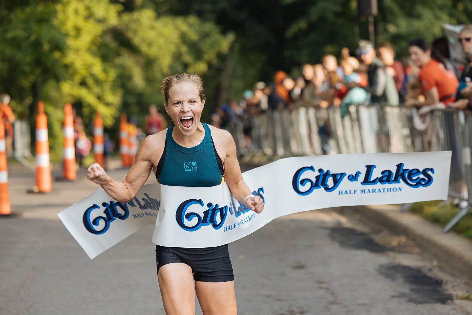 A female runner crosses the finish line with a joyful expression at the City of Lakes Half Marathon. She is wearing a blue athletic tank top and black shorts, and is breaking through the race banner.