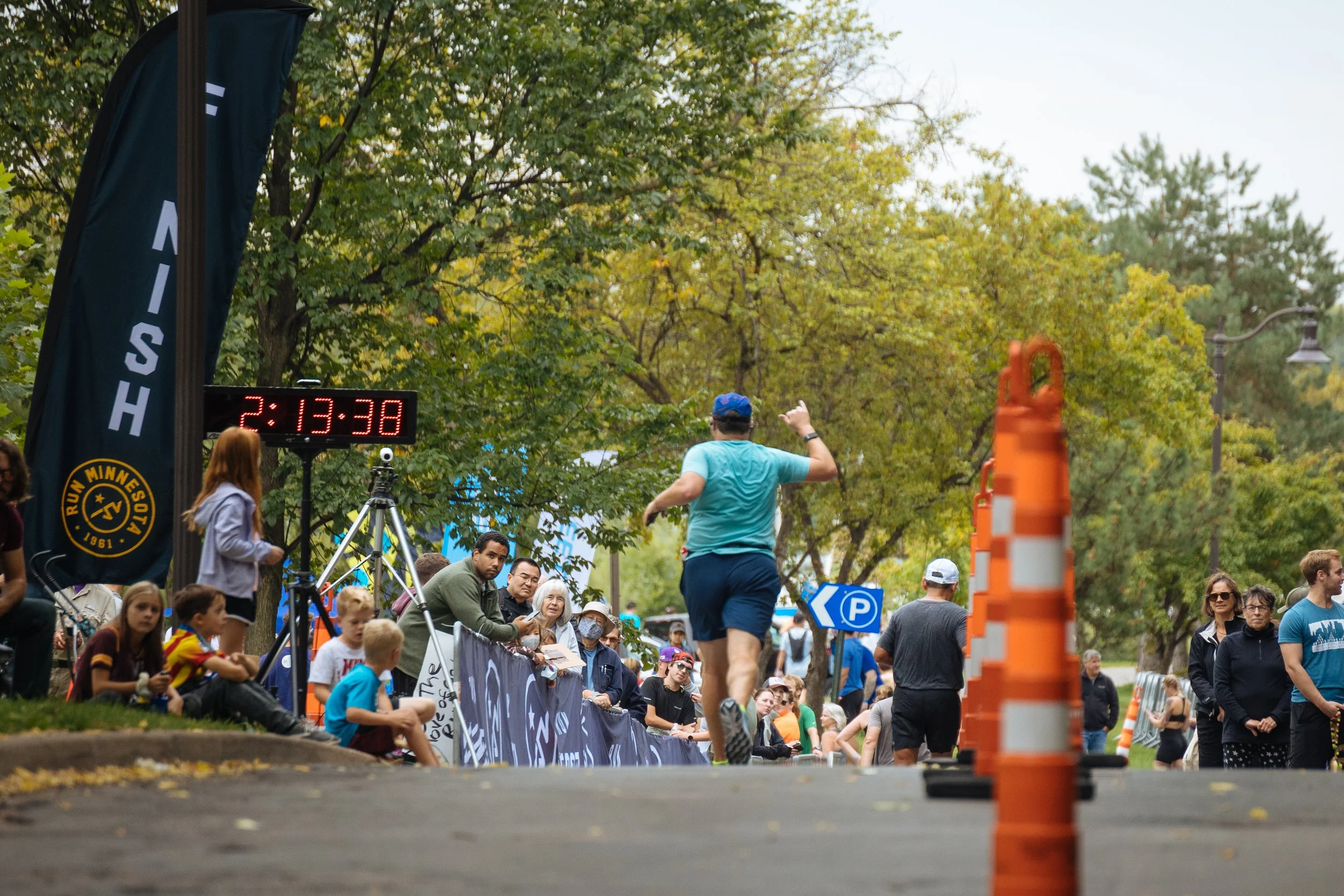 Crowd gathered at an outdoor running event, with a runner in blue approaching the finish line, surrounded by spectators and event signage, with green trees and overcast sky in the background.