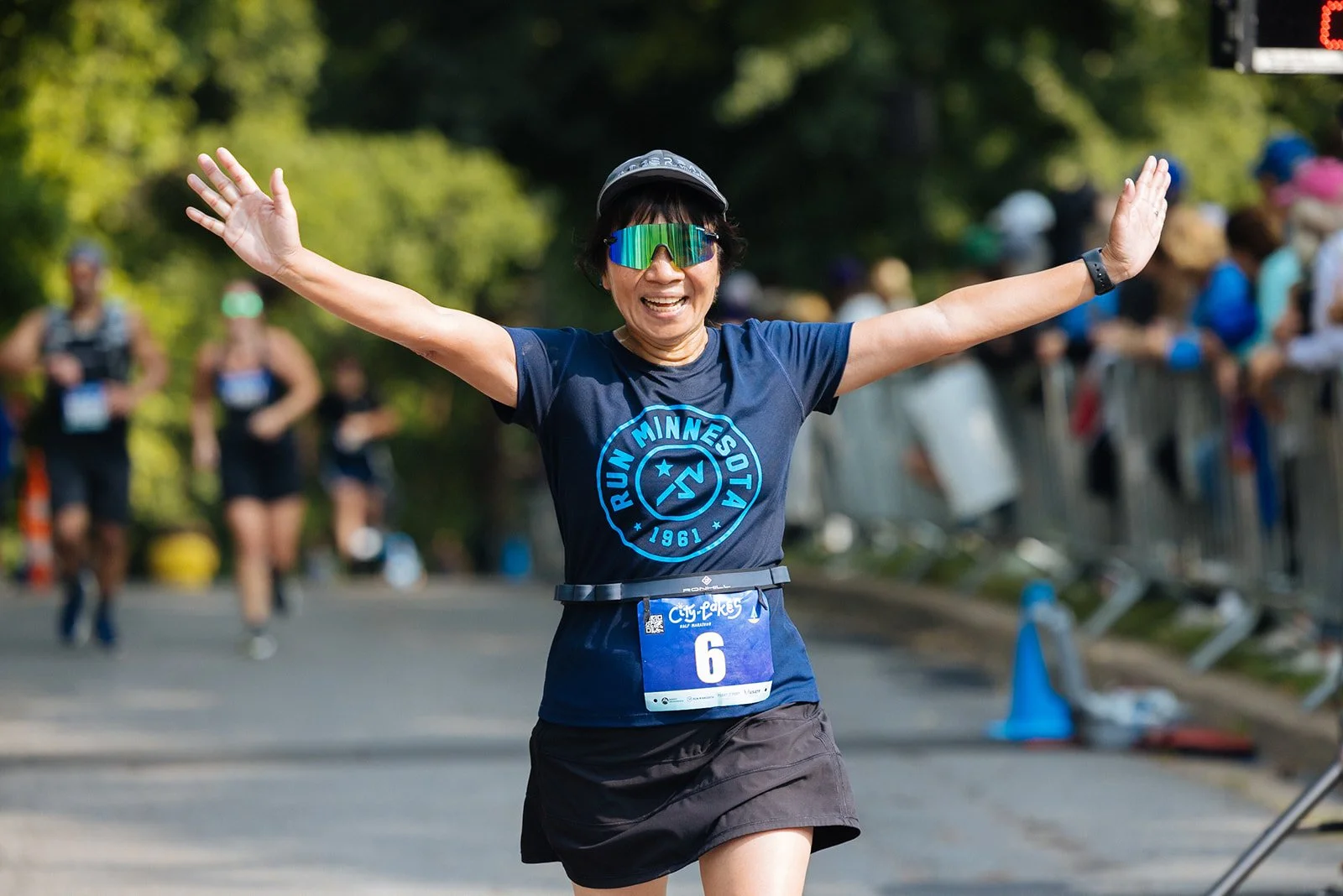 A woman in athletic gear celebrating as she crosses the finish line of a marathon, with her arms raised and a big smile on her face, wearing sunglasses and a bib number 6, in a park setting with trees and other runners in the background.