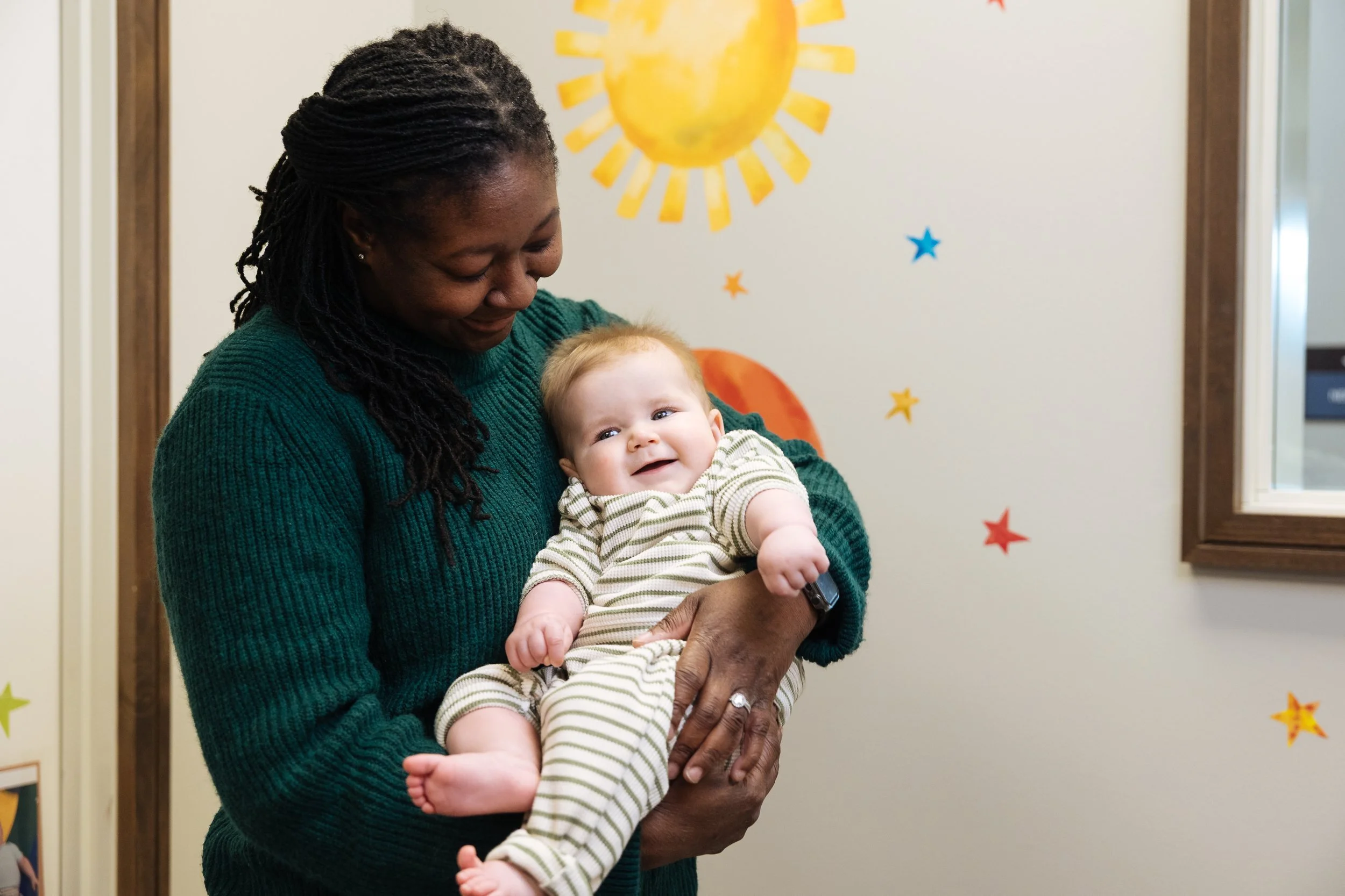 An African American woman wearing a green sweater holds a smiling baby with blonde hair dressed in a striped outfit. They are indoors against a white wall decorated with colorful star and sun-shaped wall decals.