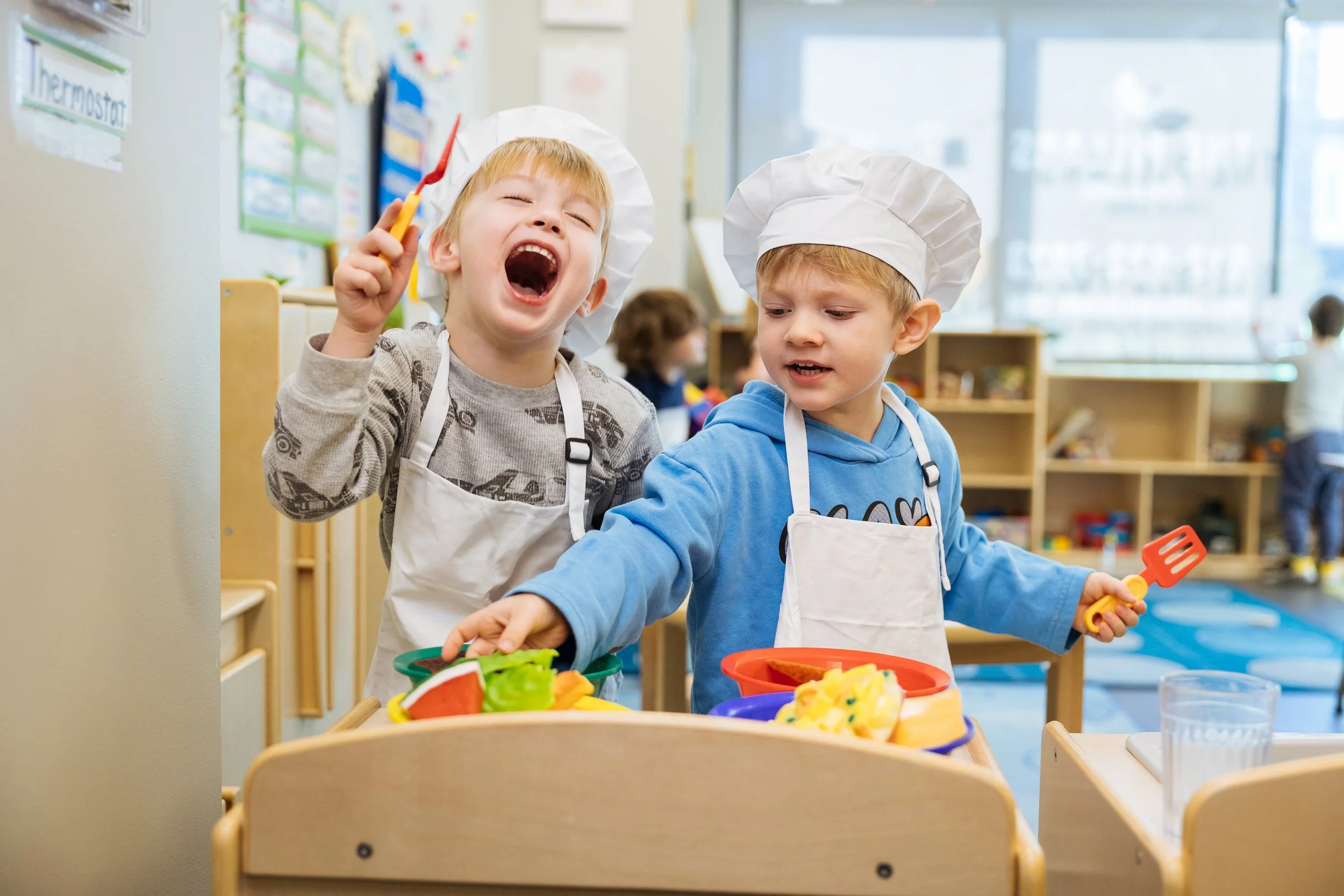 Two young boys wearing chef hats and aprons preparing food at a preschool or classroom setting. One boy is smiling and holding a yellow kitchen tool, while the other is focused on the food.