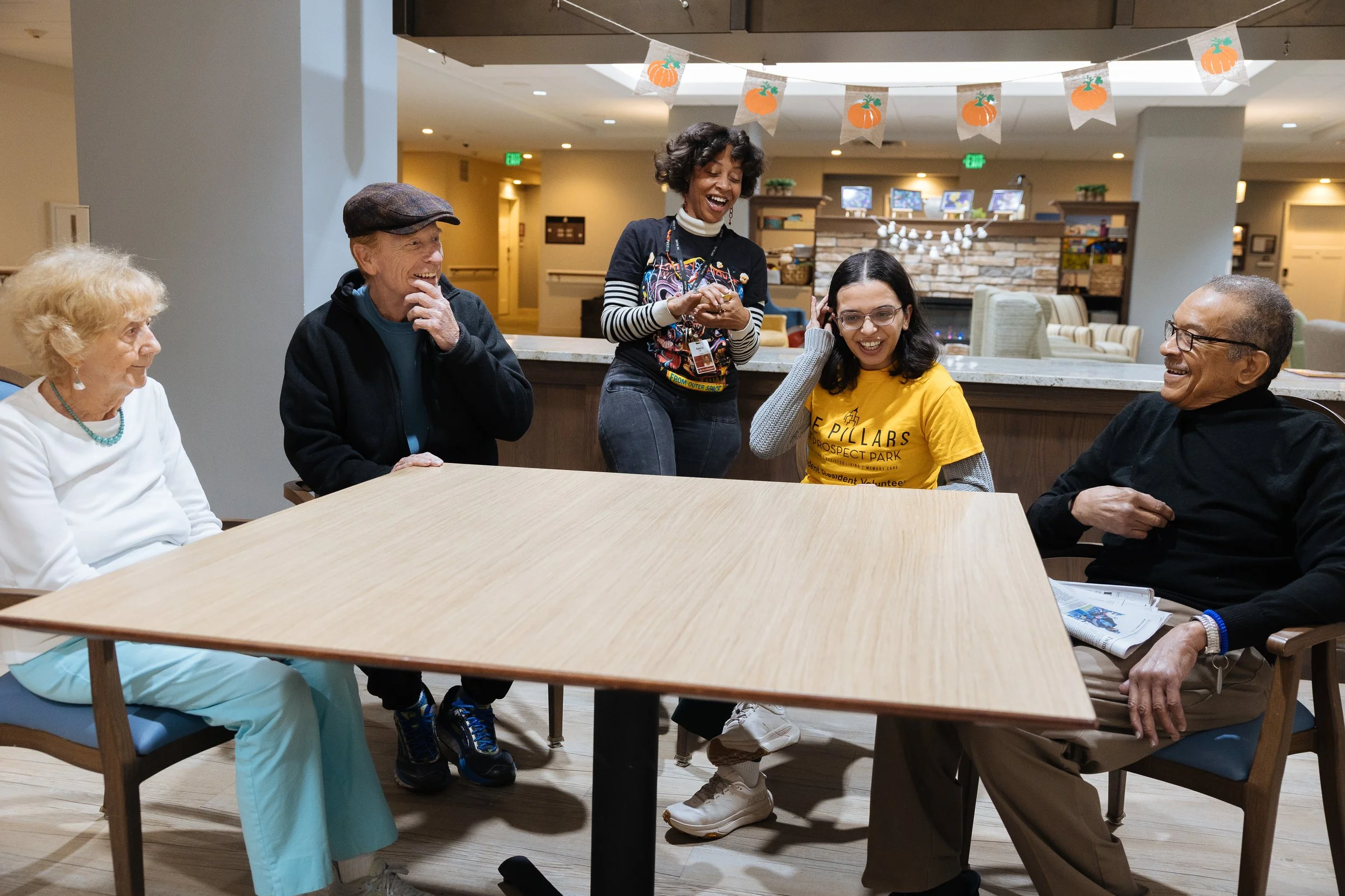 A group of five diverse elderly and middle-aged adults sitting around a table, laughing and smiling while engaging in conversation, with festive pumpkin decorations hanging above in a warmly lit, cozy indoor space.