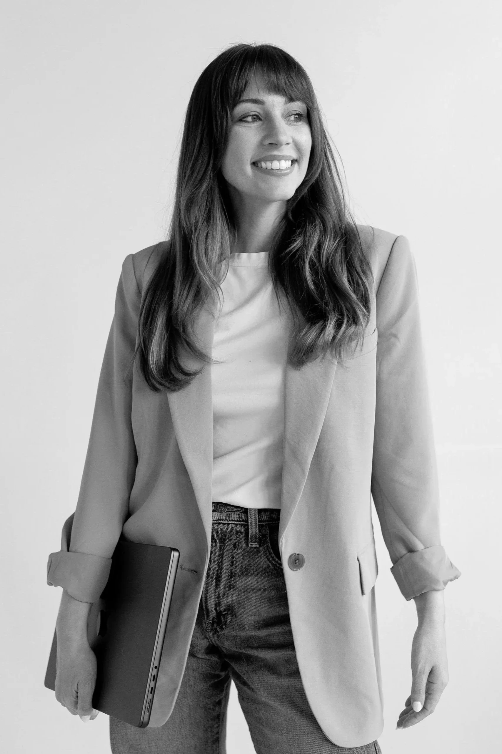 A woman in a blazer holding a closed laptop, standing against a plain background.