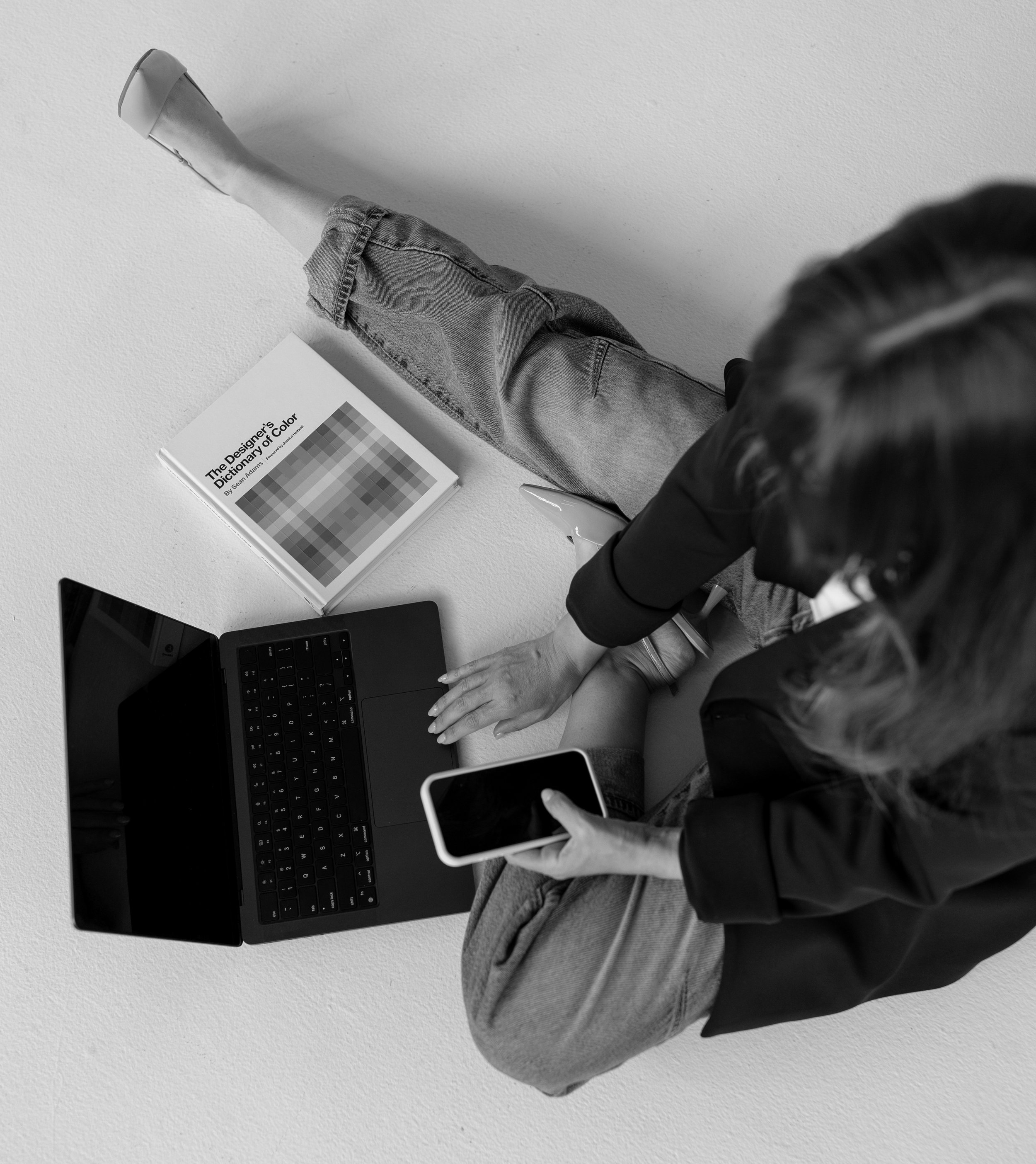 Top view of a person wearing a black blazer and gray pants, working on a laptop and holding a smartphone over a white table with a color palette book and a pen.