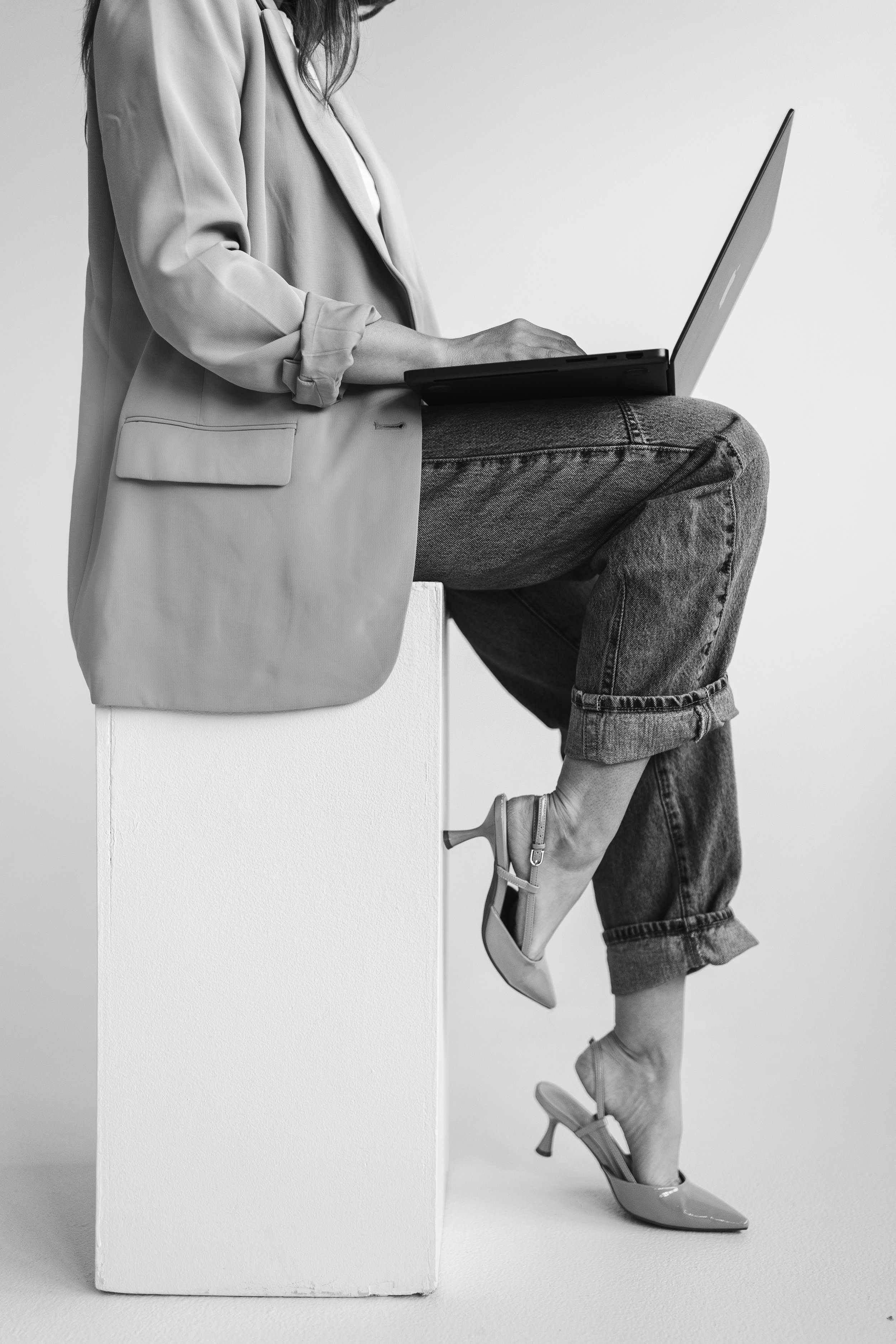 Black and white photo of a woman sitting on a high stool with her legs crossed, wearing a blazer, jeans, and high heels, with a laptop on her lap. The image shows a creative split view, with one side of her body and the stool behind a white wall.