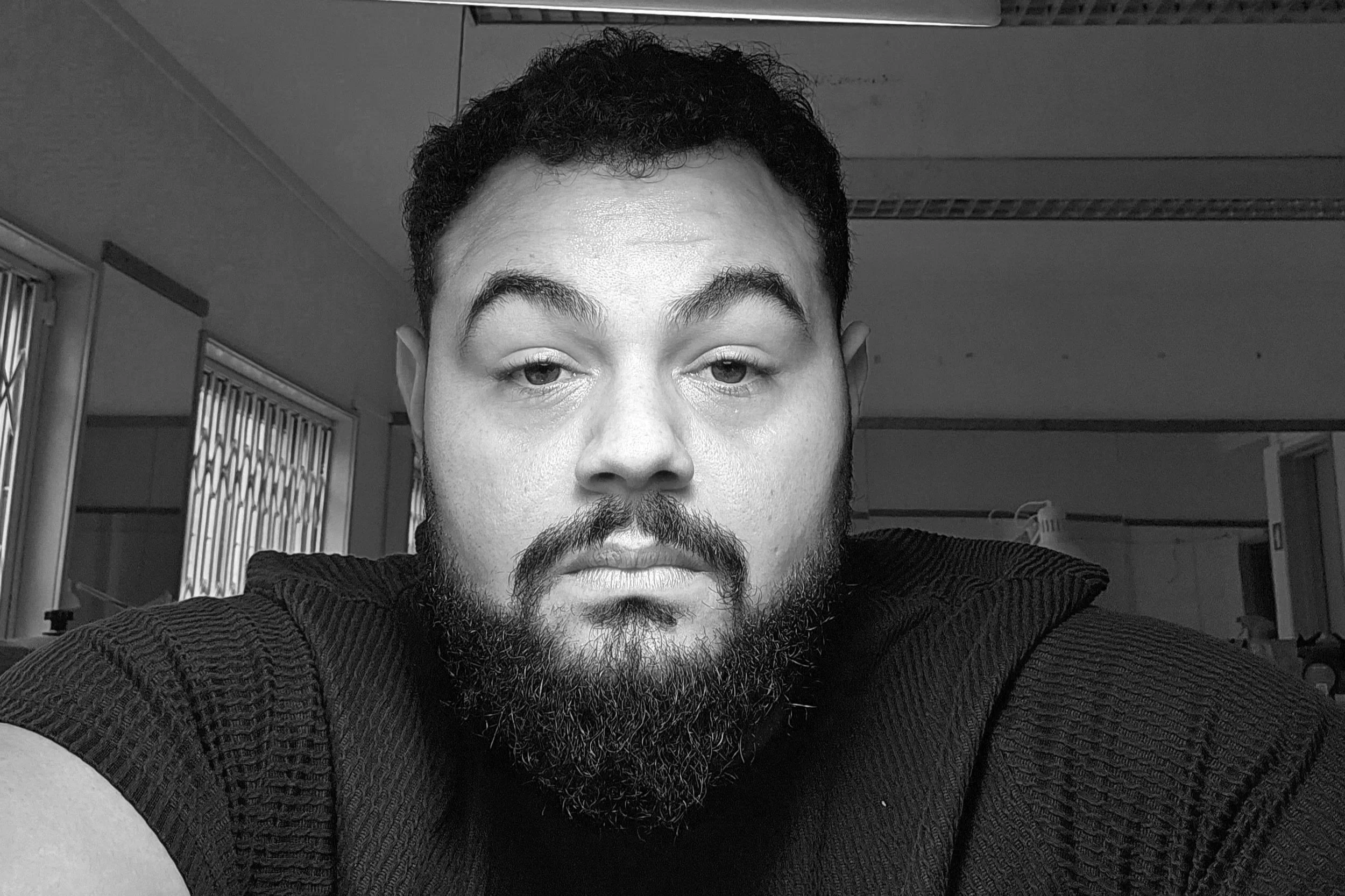 Retrato em preto e branco de um homem com barba e cabelo encaracolado curto com uma expressão séria, usando camiseta preta, em um ambiente interno.
