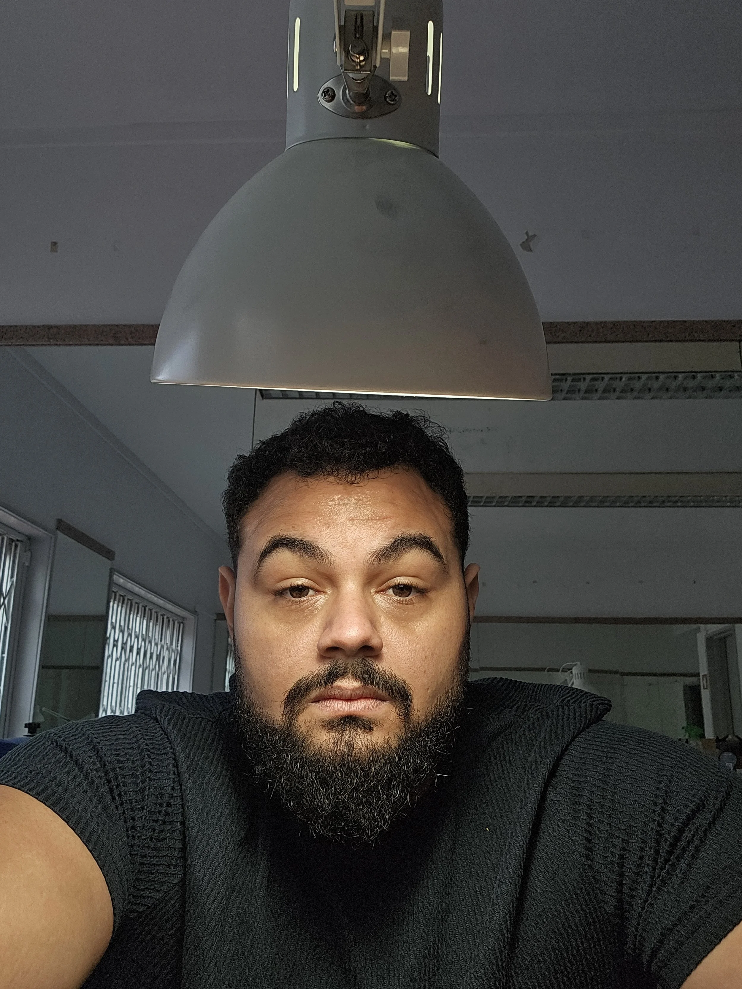 Homem com barba e cabelo encaracolado curto usando camiseta preta, em um ambiente interno com iluminação no rosto e fundo escurecido fundo.