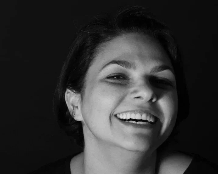 Retrato em preto e branco de uma mulher sorrindo, usando camiseta preta, com cabelo curto e fundo escuro.