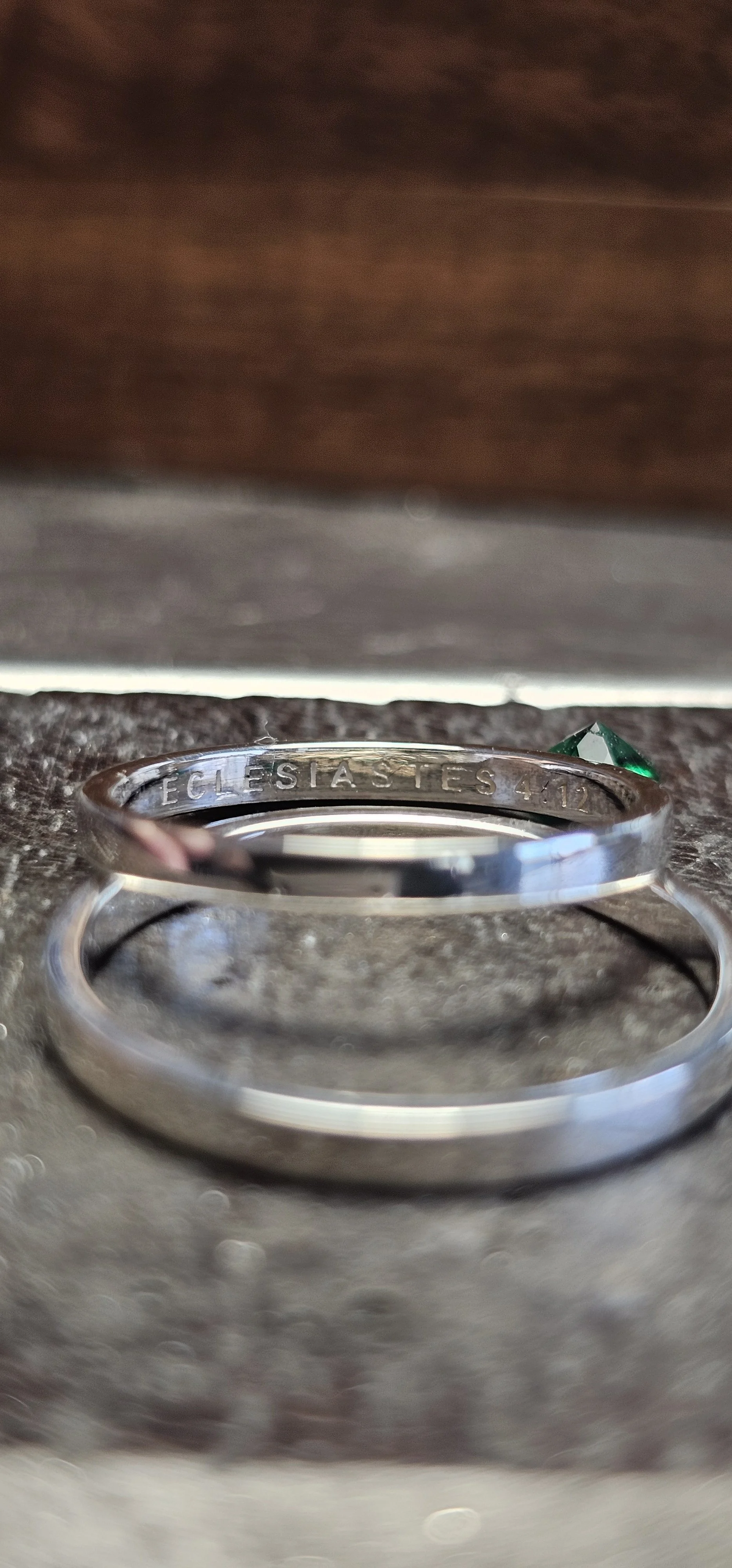 Anel de metal com inscrição "ECLESIASTES 4:12" e uma pedra verde ao lado, posicionado sobre uma superfície de pedra.