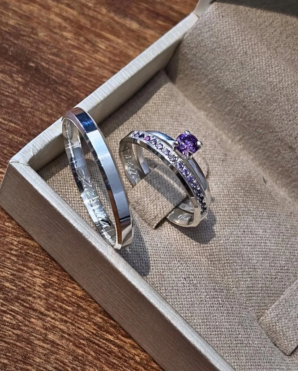 Conjunto de anéis de prata, com o da direita contendo detalhes compostos por uma pedra preciosa roxa, dentro de joias de tecido.