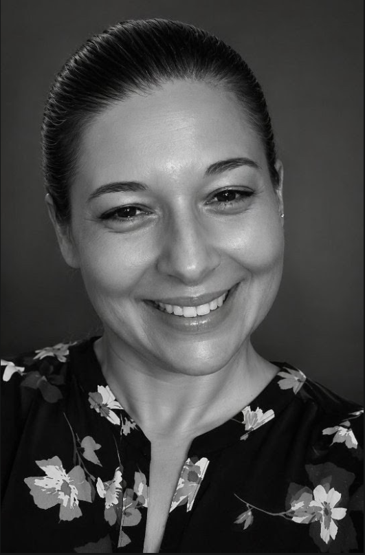 Retrato em preto e branco de uma mulher sorridente com cabelo preso, vestindo blusa com estampa de flores, olhando para a câmera.