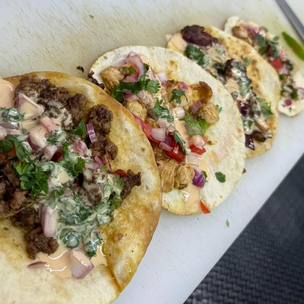 Three tacos filled with seasoned ground beef, grilled chicken, and a mixture of chopped vegetables, topped with cilantro, onions, and drizzled with creamy sauces.