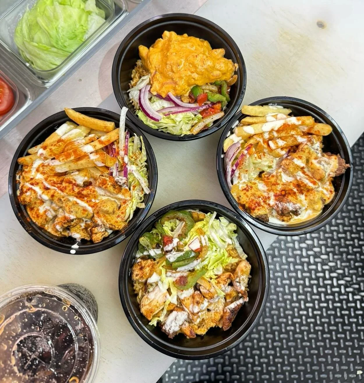 Four black bowls with chicken fajitas topped with cheese and sour cream, garnished with lettuce, onions, and peppers, on a white table. Also, a container of soda and a salad with lettuce, tomato, and cucumbers are partially visible.