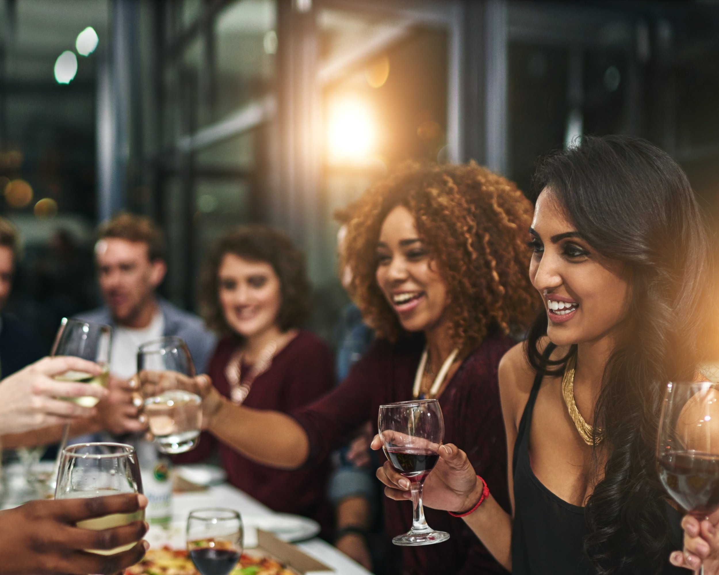 People celebrating with glasses of red wine at a party or social gathering, with a blurred background of people and a warm light.