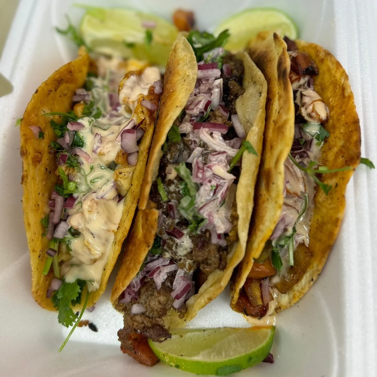 Four tacos with meat, chopped red onion, cilantro, and various sauces, garnished with lime wedges, on a white foam plate.