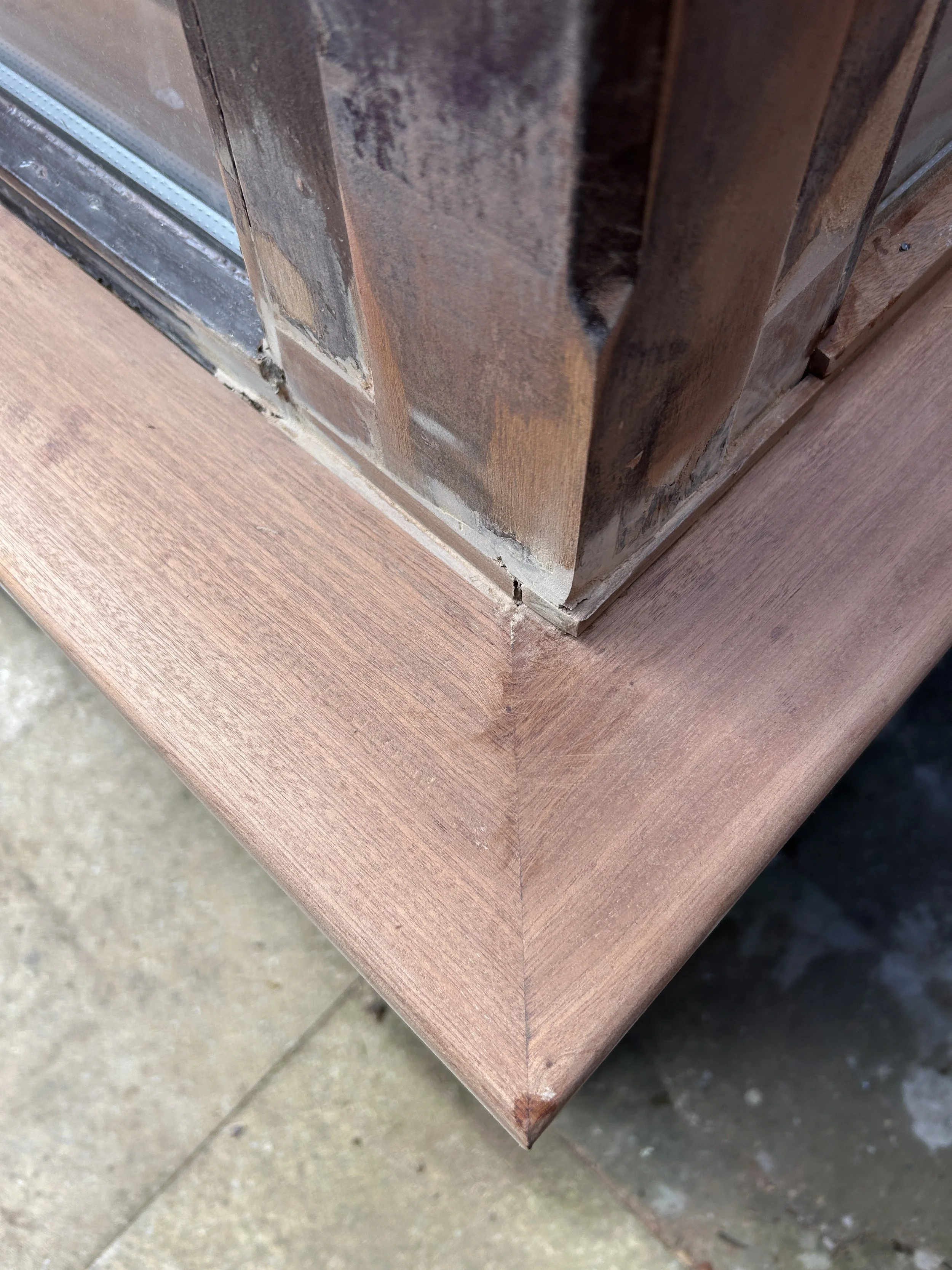 Close-up of a corner of a wooden cabinet with a glass panel, showing chipped paint and weathered wood on a concrete floor.