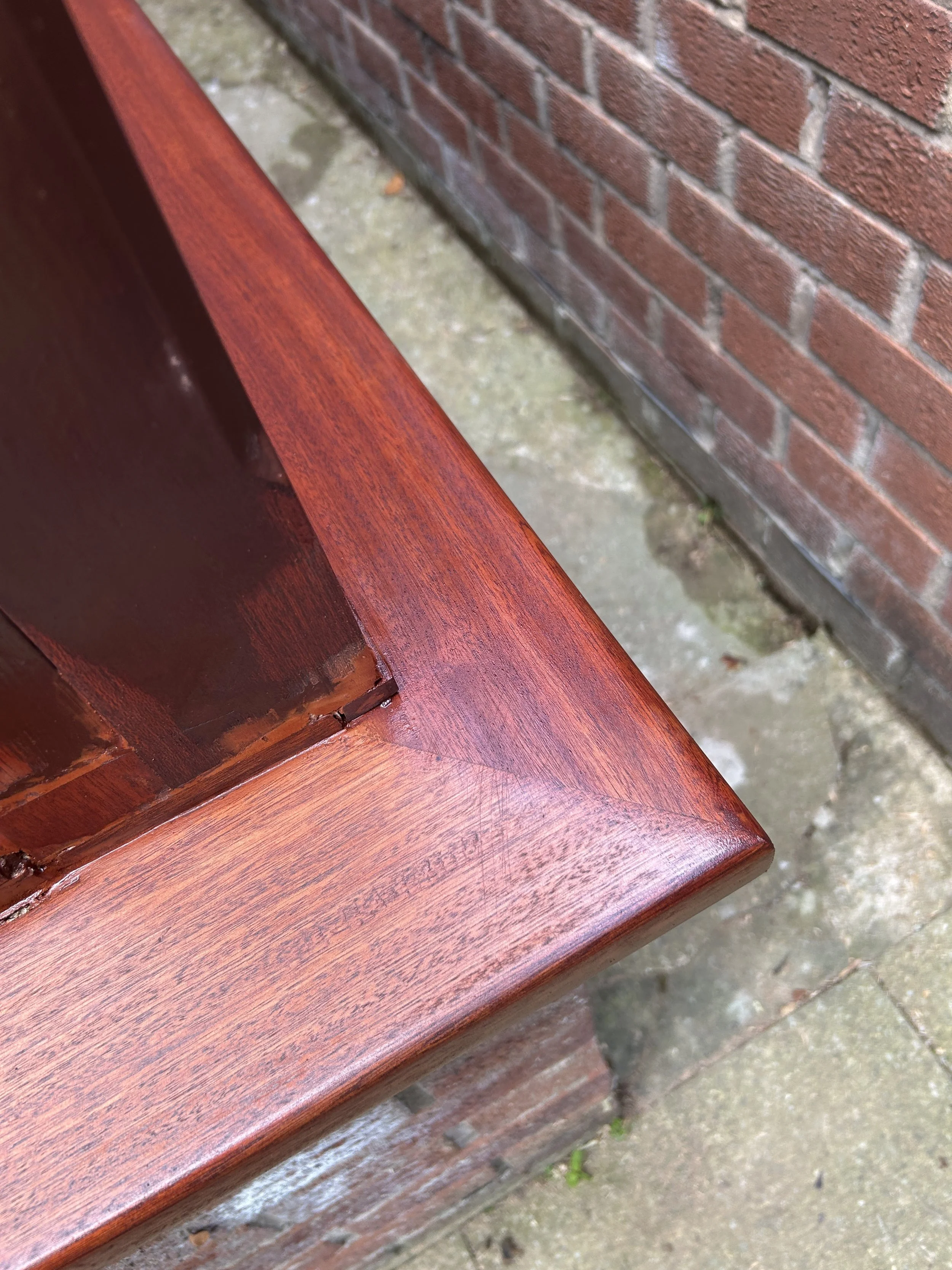 Close-up of a polished wooden bench or table corner with a brick wall and concrete ground in the background.