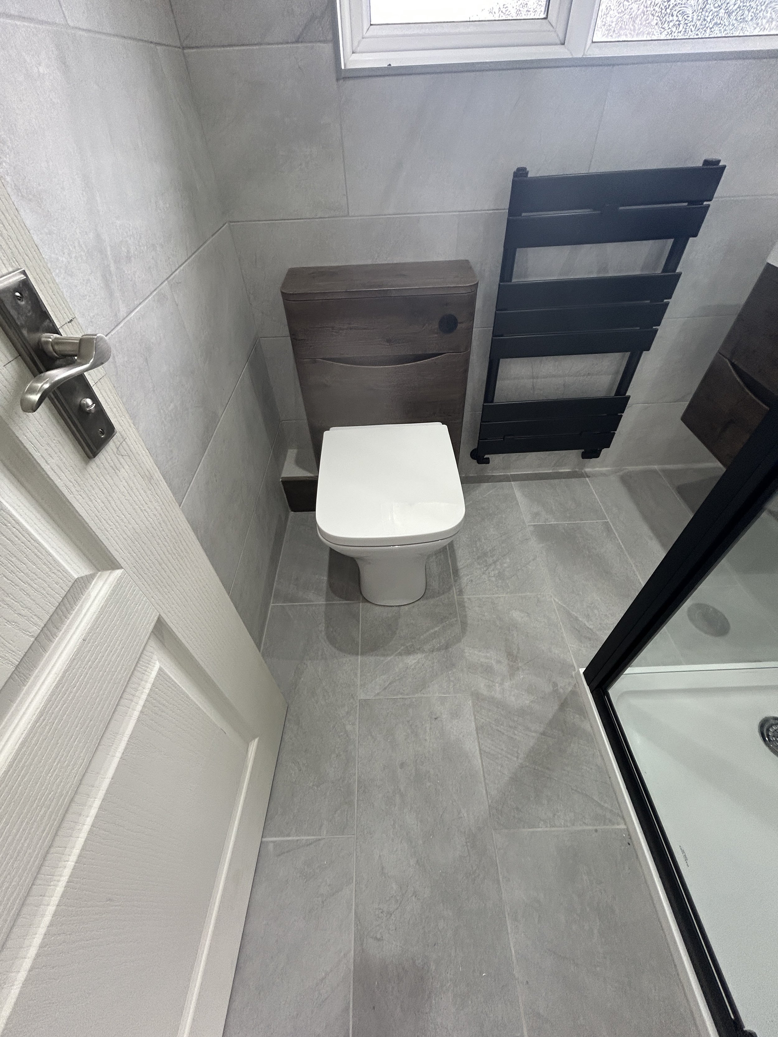 A bathroom featuring a white toilet, a wooden storage cabinet, a black towel rack, a shower door, and a window with frosted glass.
