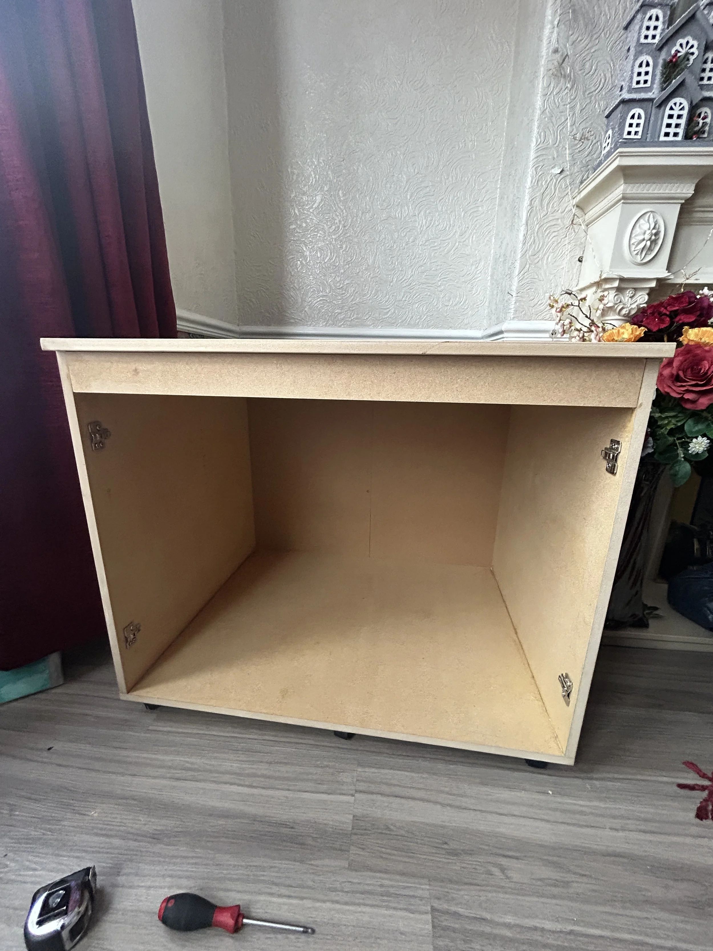 An empty wooden cabinet lying on the floor in front of a wall with textured wallpaper. There are tools, including a cordless drill, a screwdriver, and a tape measure, placed on the floor in front of the cabinet. To the right, there are decorative ite
