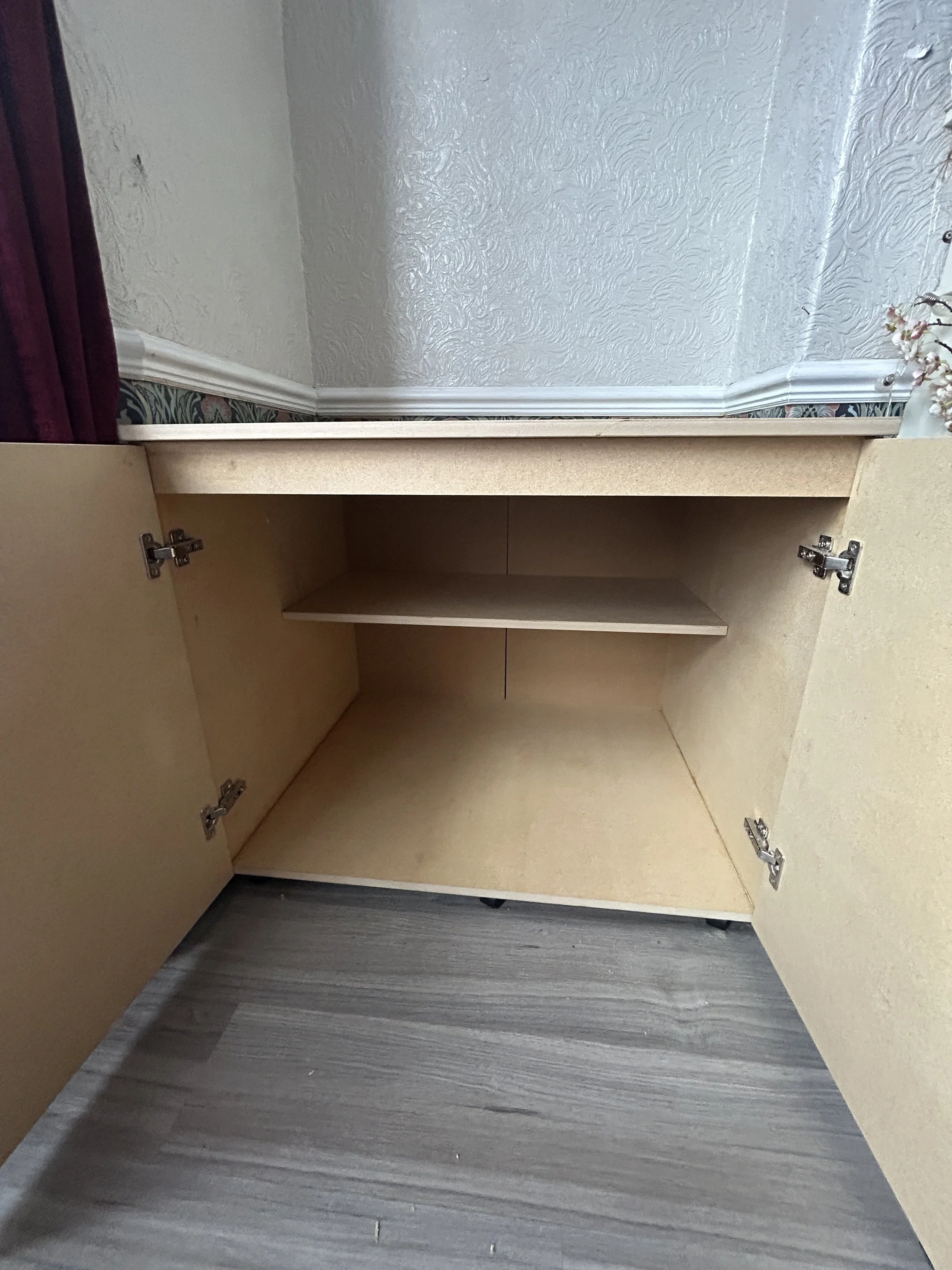 An open beige wooden cabinet with three empty shelves, mounted against a wall with textured pale blue wallpaper, a hardwood floor, and a small portion of a decorative plant on the right.