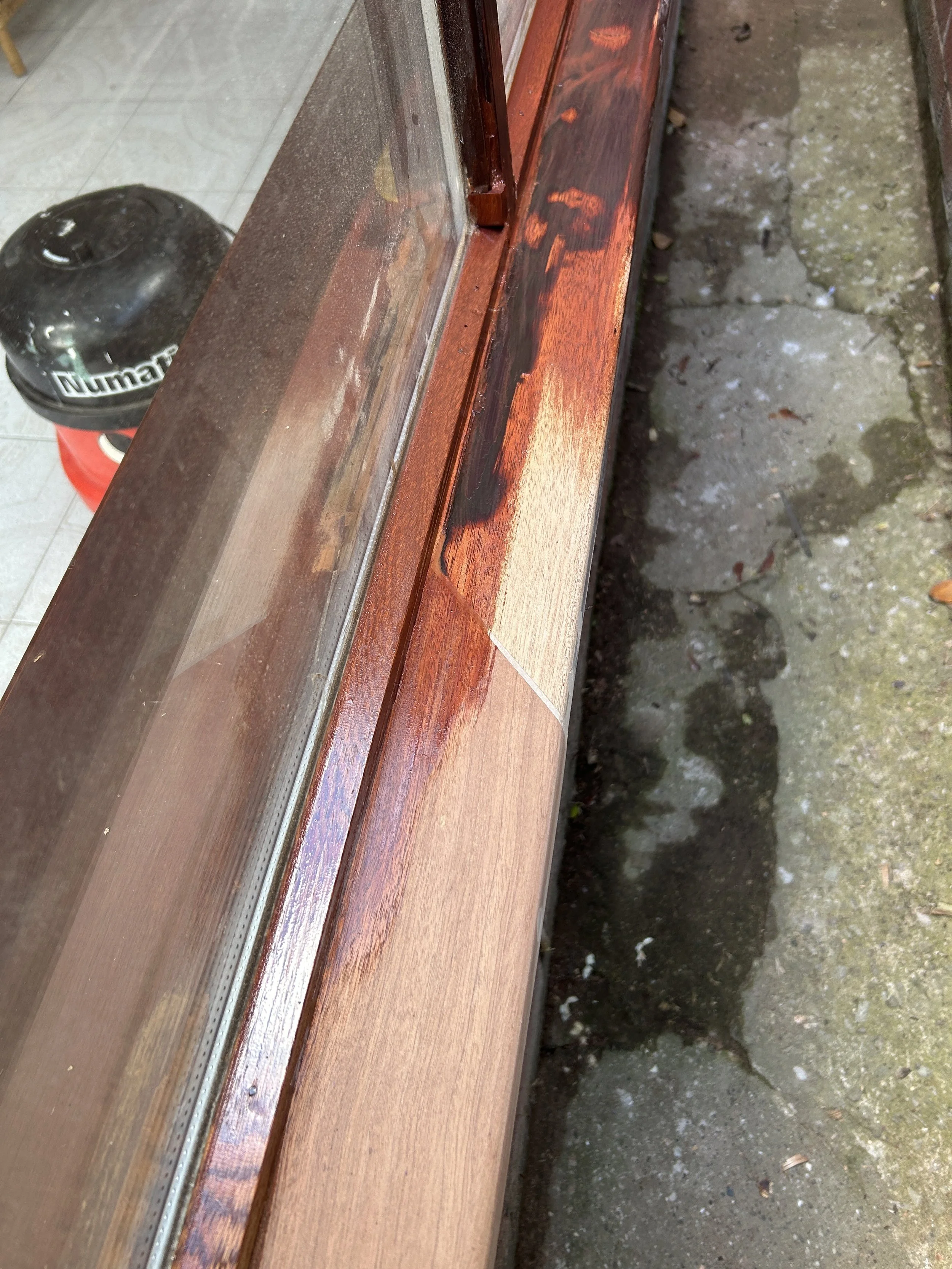 Close-up of a wooden window frame with a glass pane, showing some water spots and minor paint scratches, and a section of concrete ground outside.