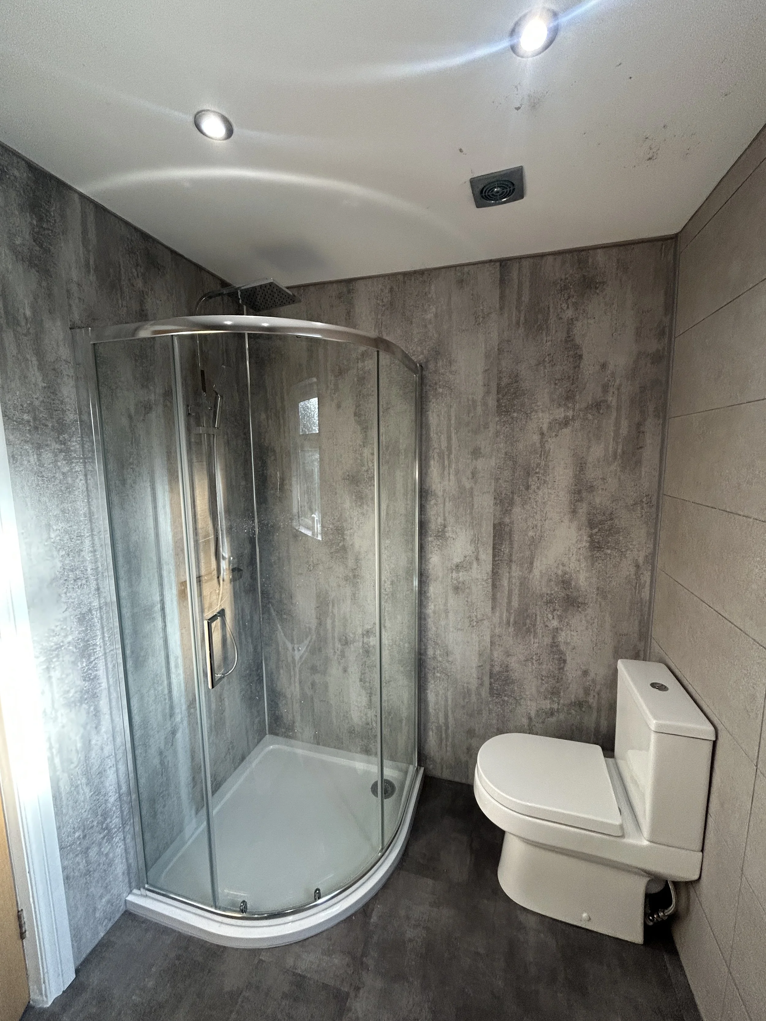 A small modern bathroom with a corner shower stall with a glass door, a rain showerhead, and a white toilet against a dark tiled floor and textured gray wall.