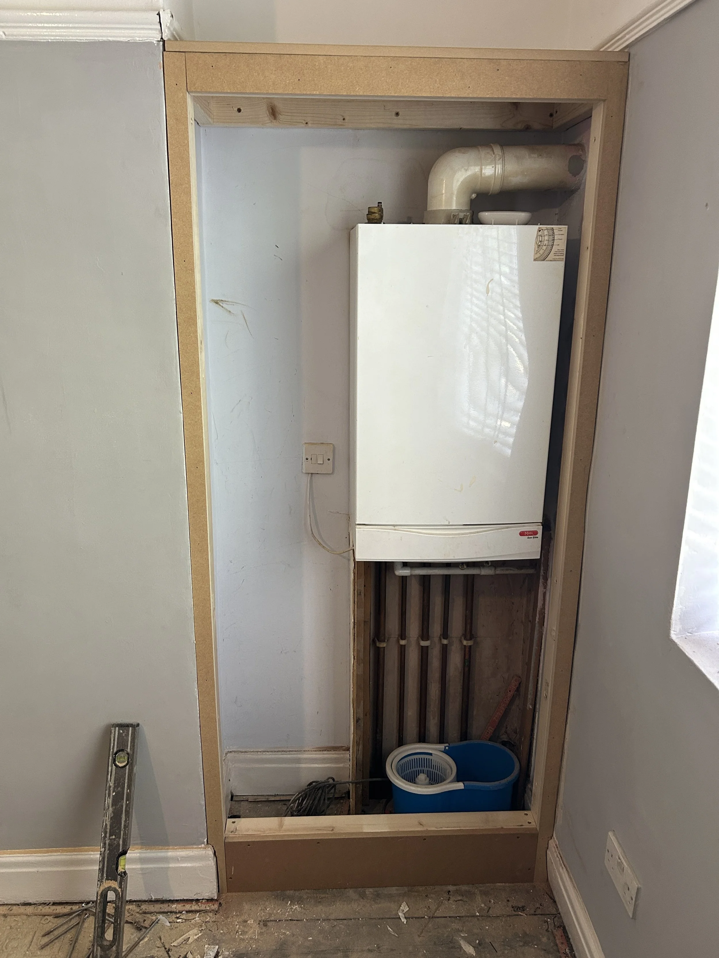 Bathroom under renovation with a small wooden frame around a space for a washing machine. A washing machine is installed inside the frame, visible from the side. There is a blue laundry basket with a laundry crisper inside, a level tool on the left, 
