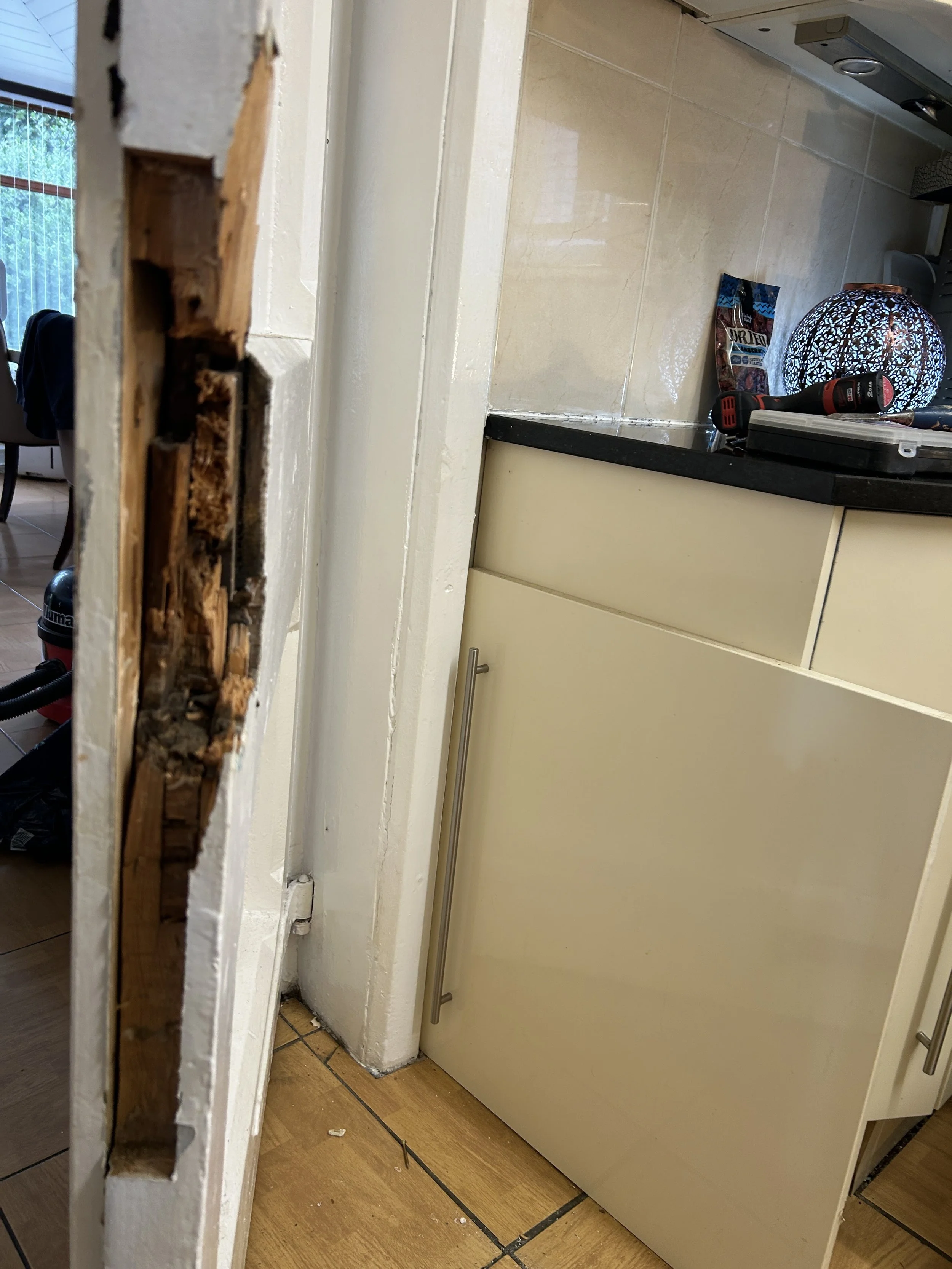 Close-up of a damaged door frame and a kitchen and tiled floor visible in the background with various items including a lamp, drill, and a bag on the kitchen counter.
