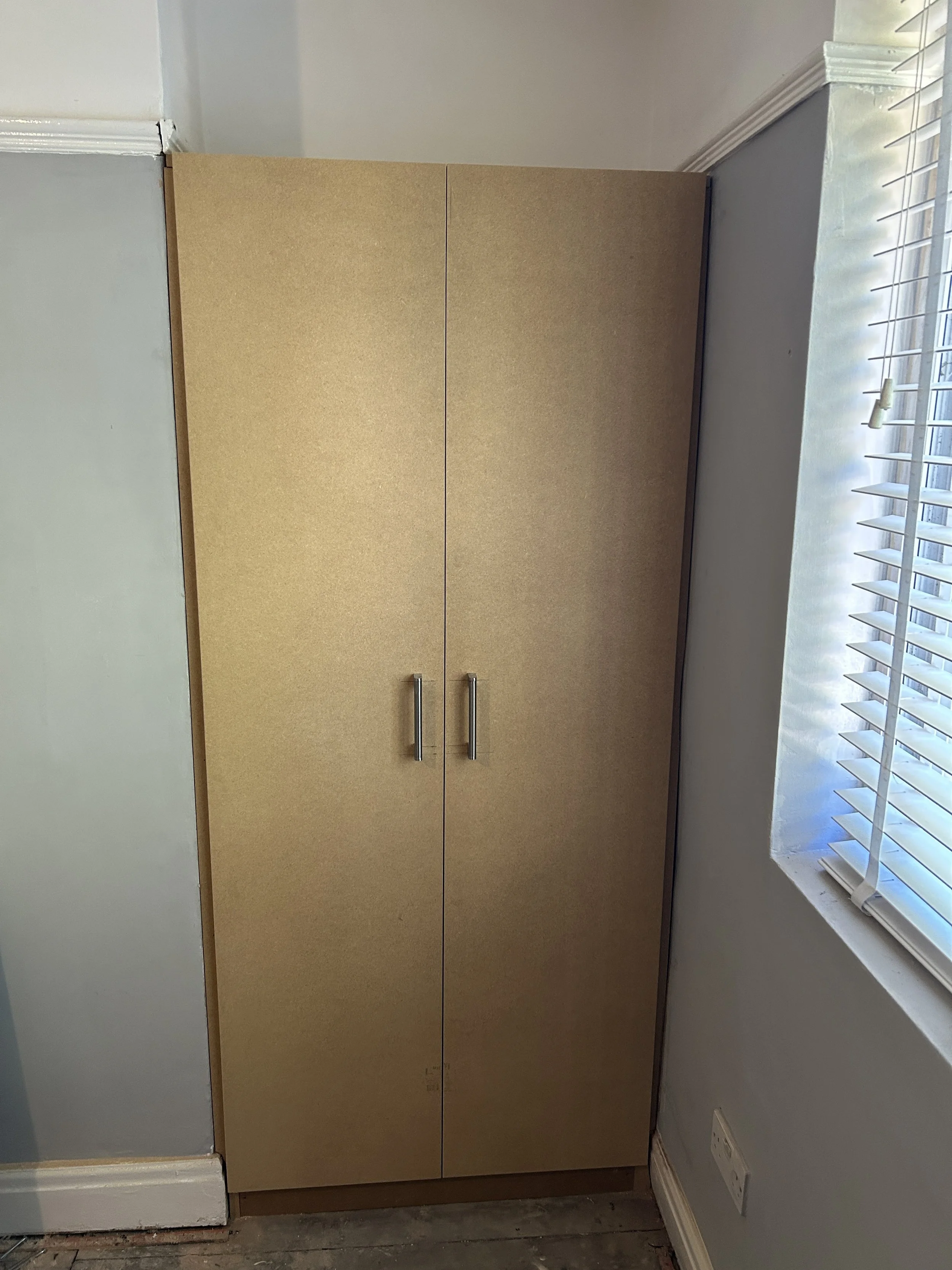 A beige kitchen cabinet with two doors and metal handles, positioned next to a window with white blinds in a room with light blue walls and white trim.