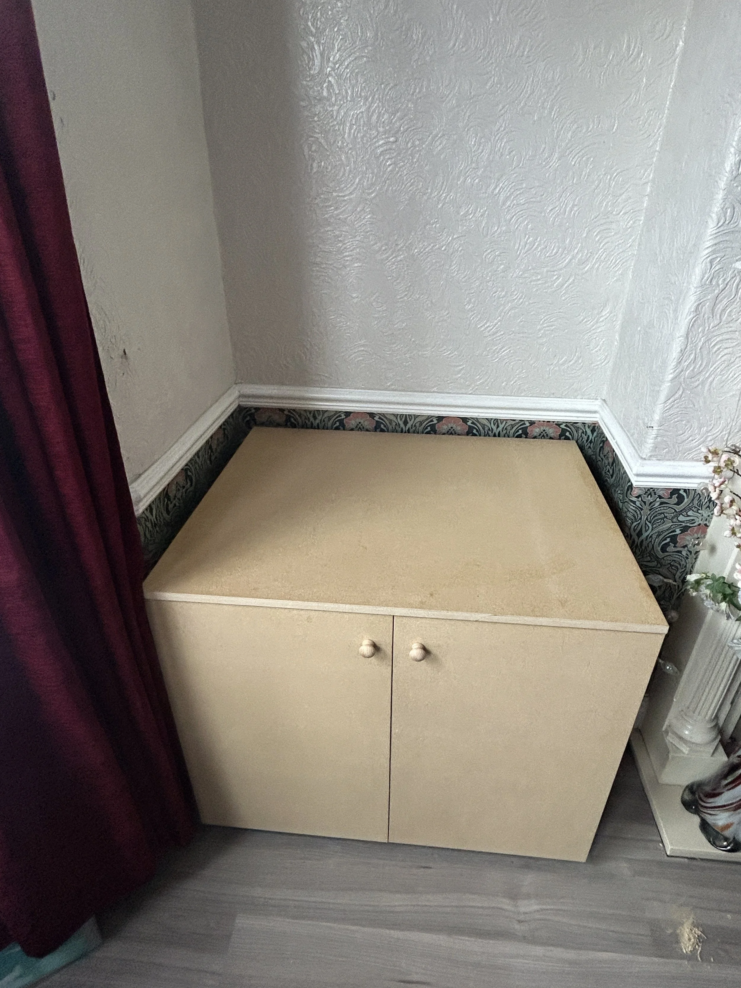 A small light-colored cabinet with two doors and round knobs, positioned against a wall with textured white wallpaper and decorative border at the bottom. Part of a dark red curtain is visible on the left side, and the floor is wood-like.