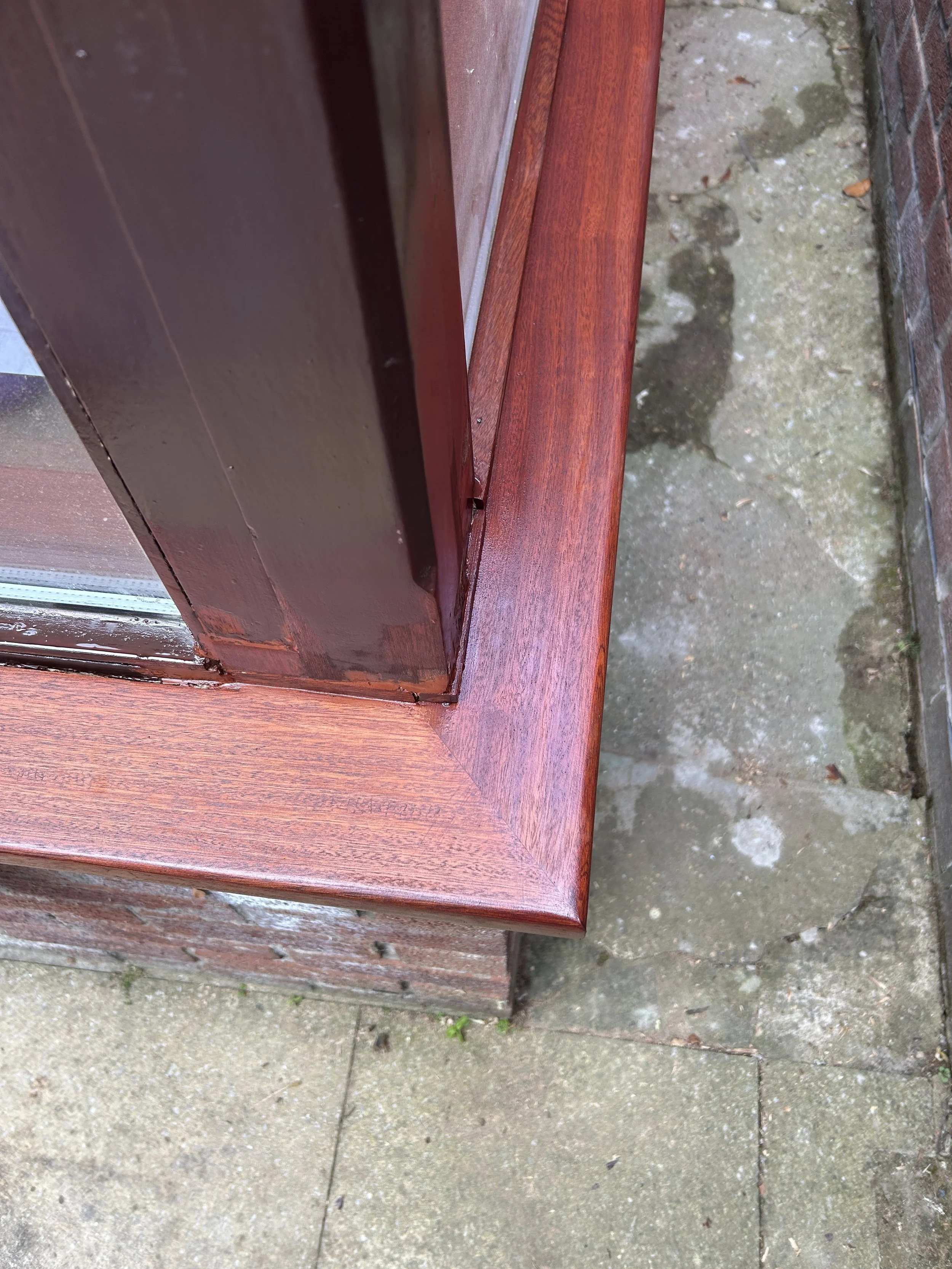 Close-up of a wooden window frame, showing its reddish-brown finish, with a concrete and brick exterior wall nearby.