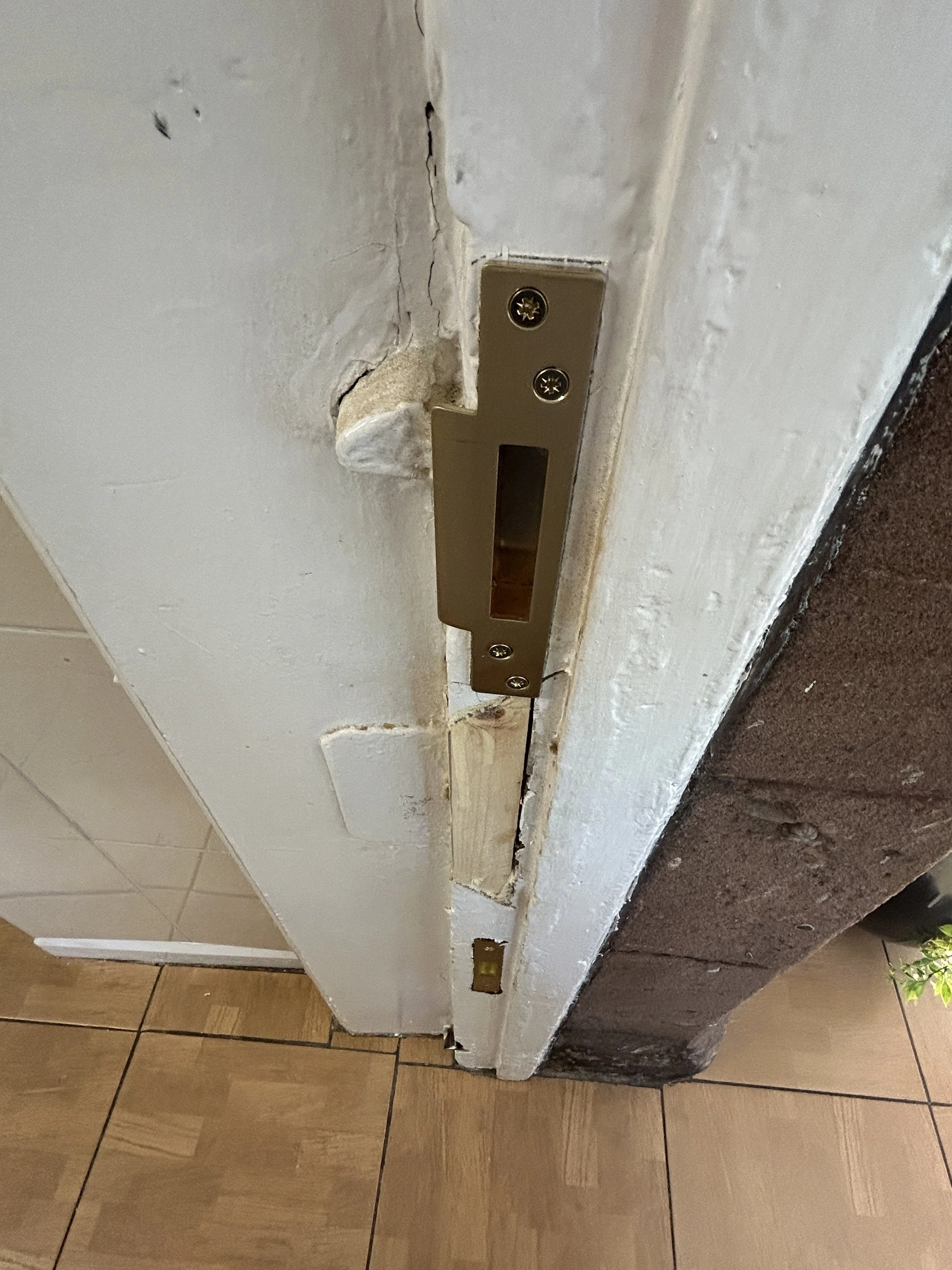 Close-up of a door's bottom edge showing a metal strike plate and surrounding damage to the door frame and wall.