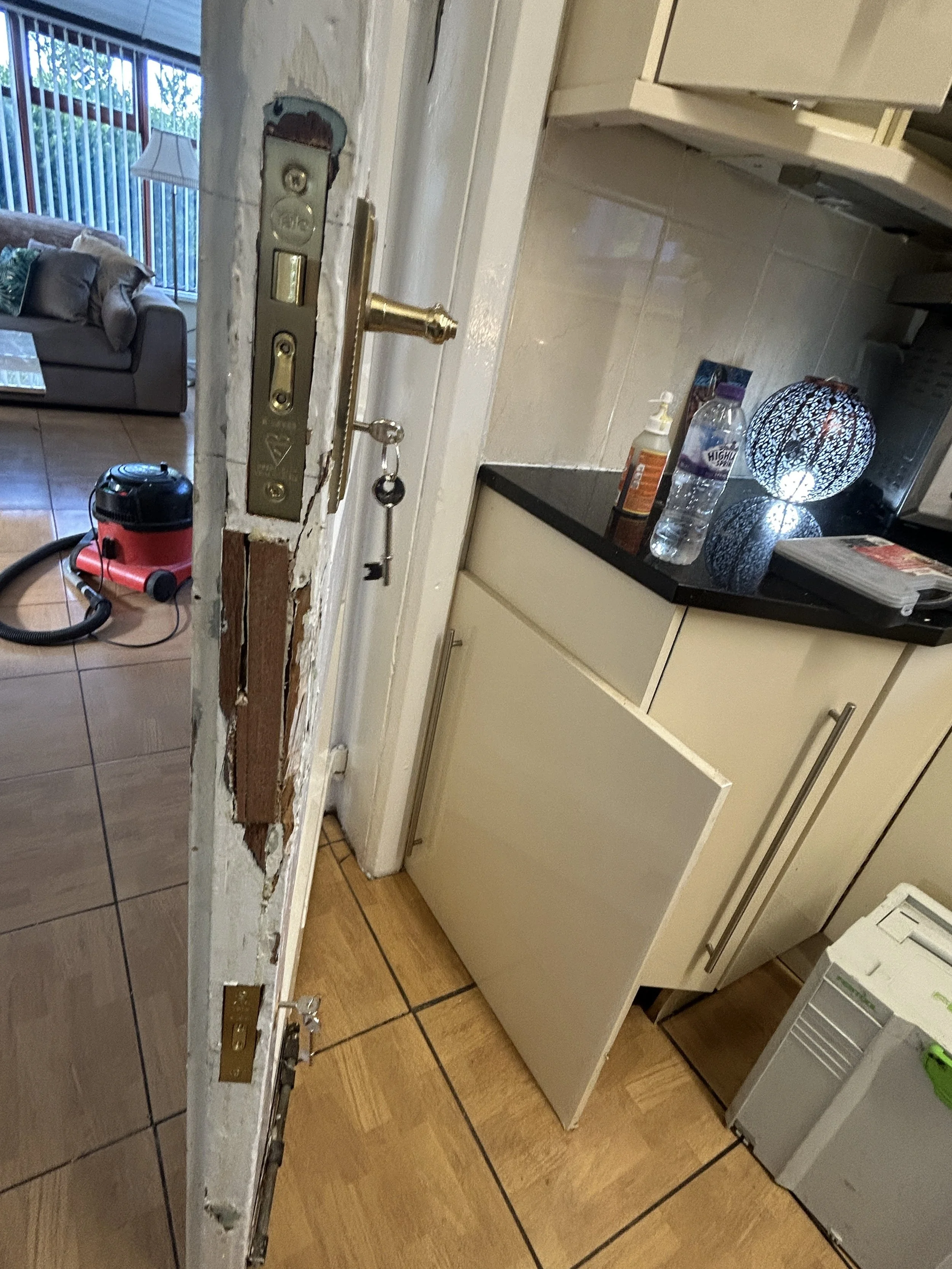 The photo shows a kitchen counter with a partially removed section, exposing the wall and electrical outlets. There is a door knob and key hanging from the wall. In the background, a living room with a sofa, window, and a vacuum cleaner on the floor 