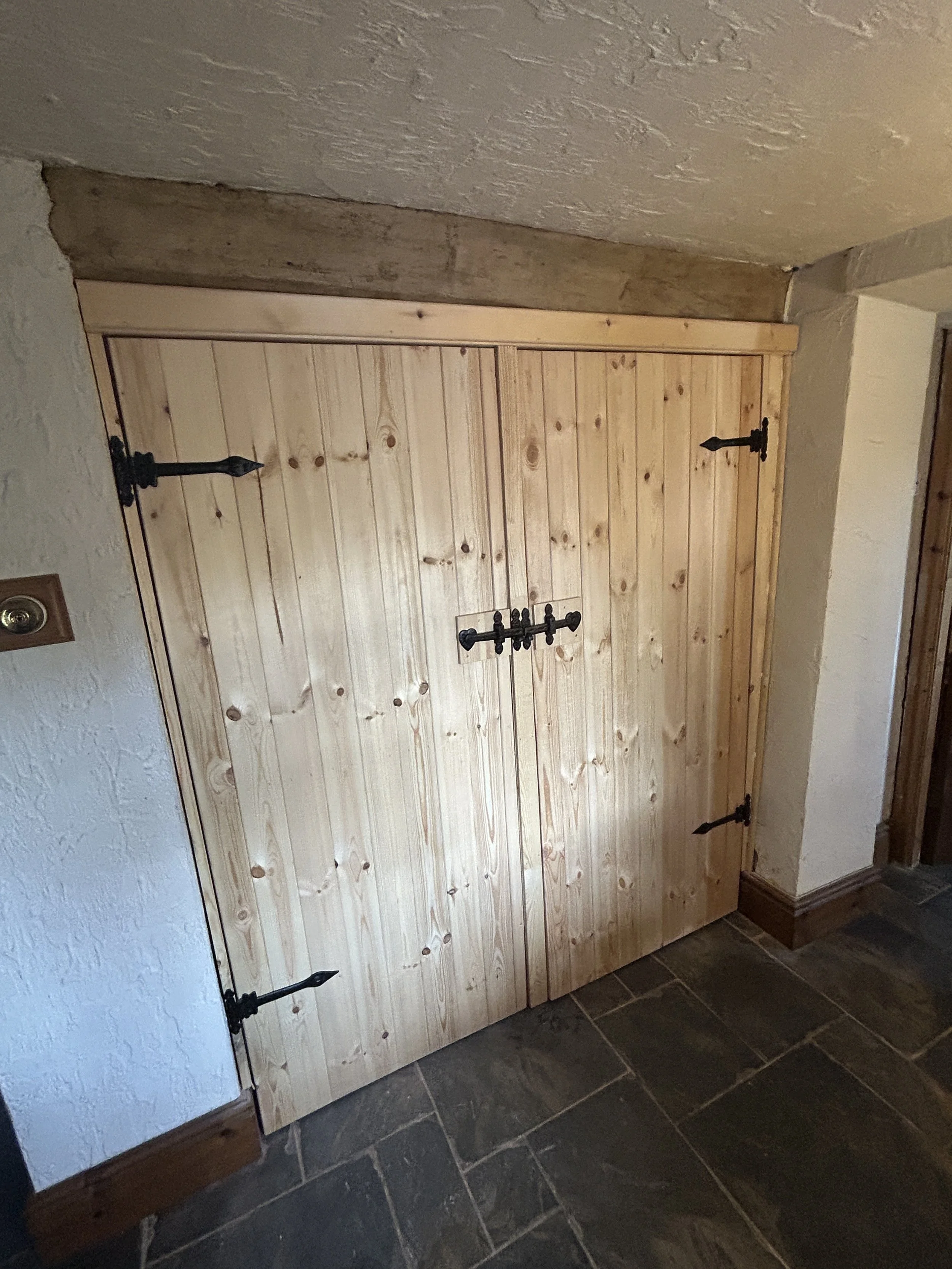 A wooden closet door with black metal hinges and a latch, set in a rustic interior with stone flooring and textured white walls.