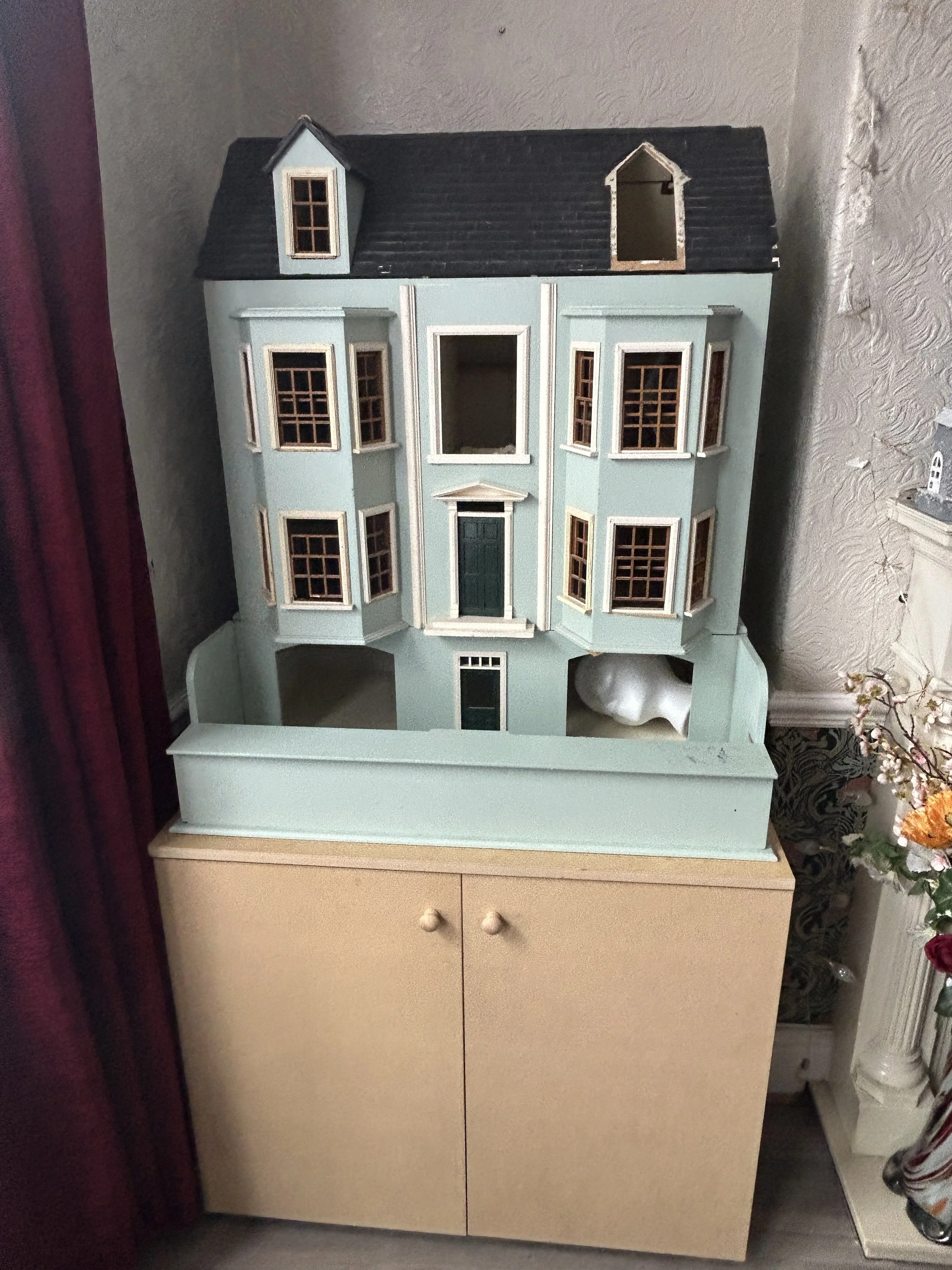 A miniature dollhouse painted in light blue with white trim, featuring multiple windows and a central front door, placed on a wooden piece of furniture.