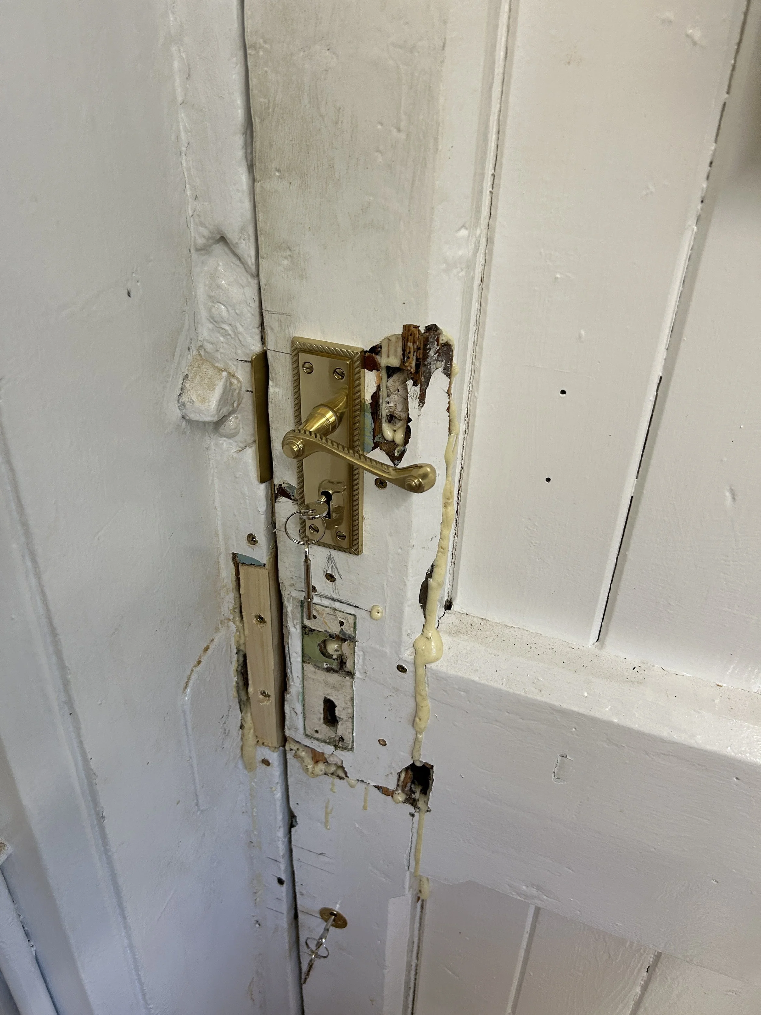 A broken door lock with exposed and damaged components, with the door painted white and showing signs of wear.
