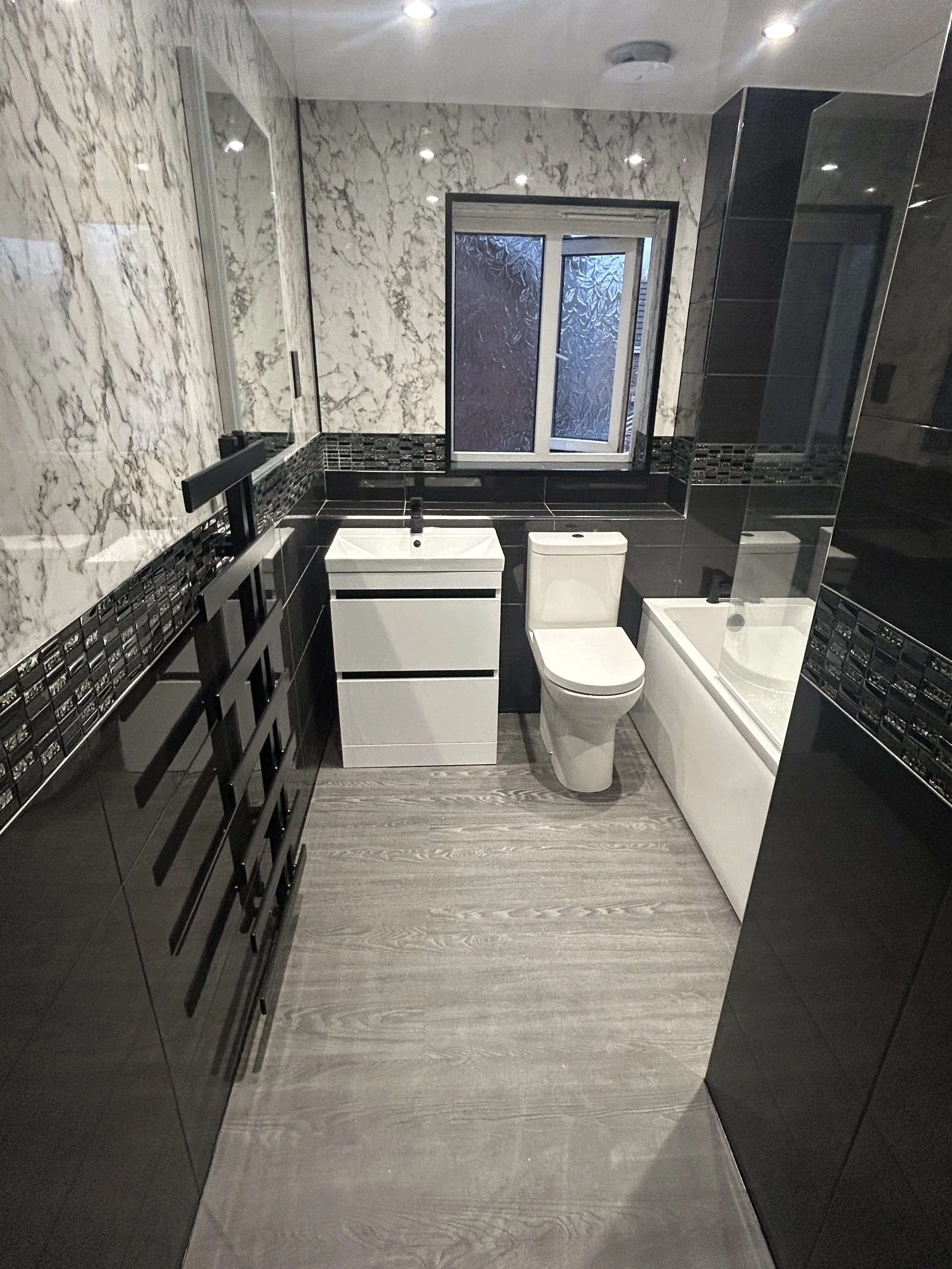 Modern bathroom with black and white marble walls, black tiled accents, vanity with white sink, toilet, shower with frosted glass window, and gray wood-look flooring.