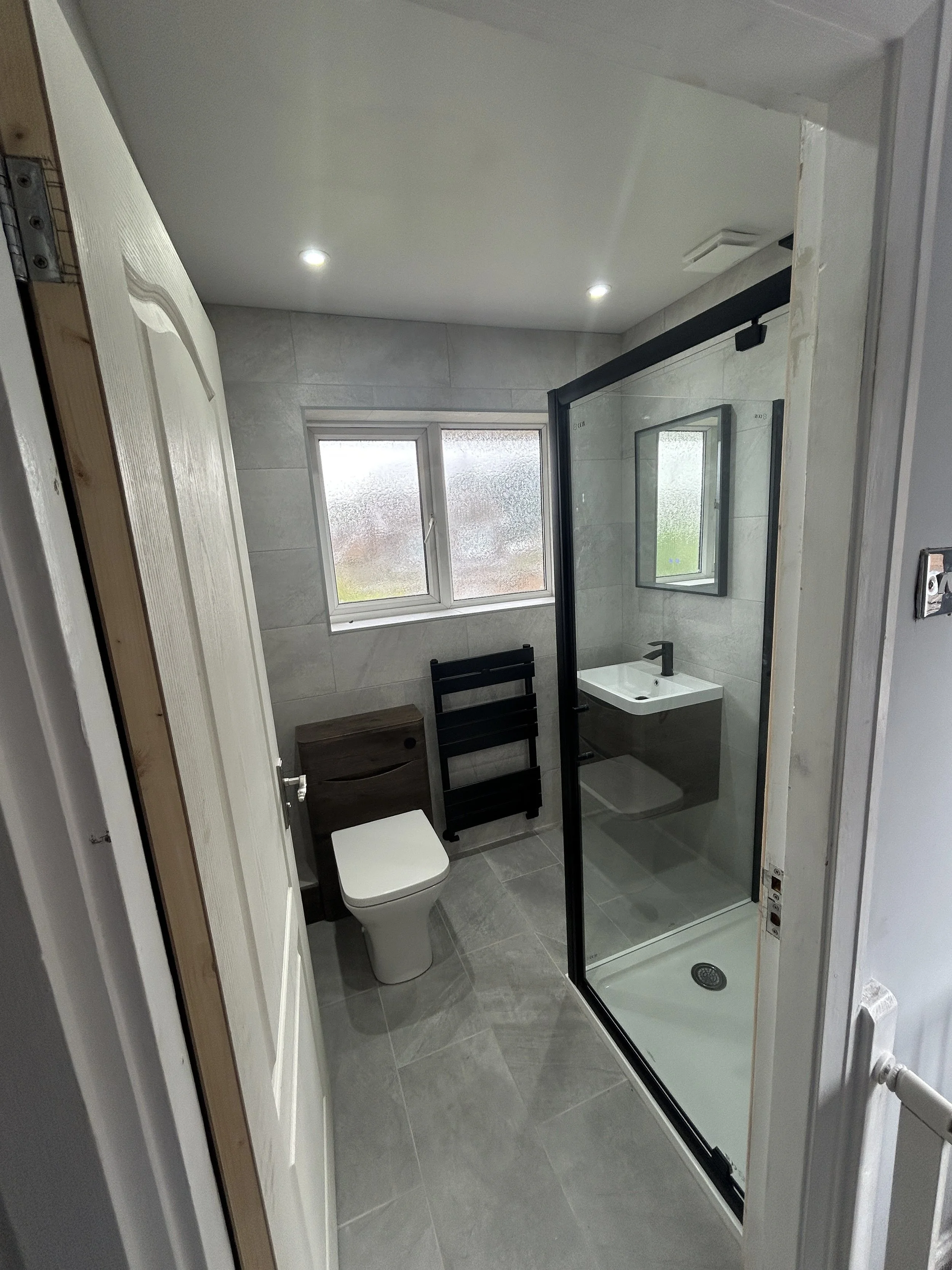 Modern bathroom with frosted window, toilet, small sink, black towel rack, and glass shower enclosure with black frame.