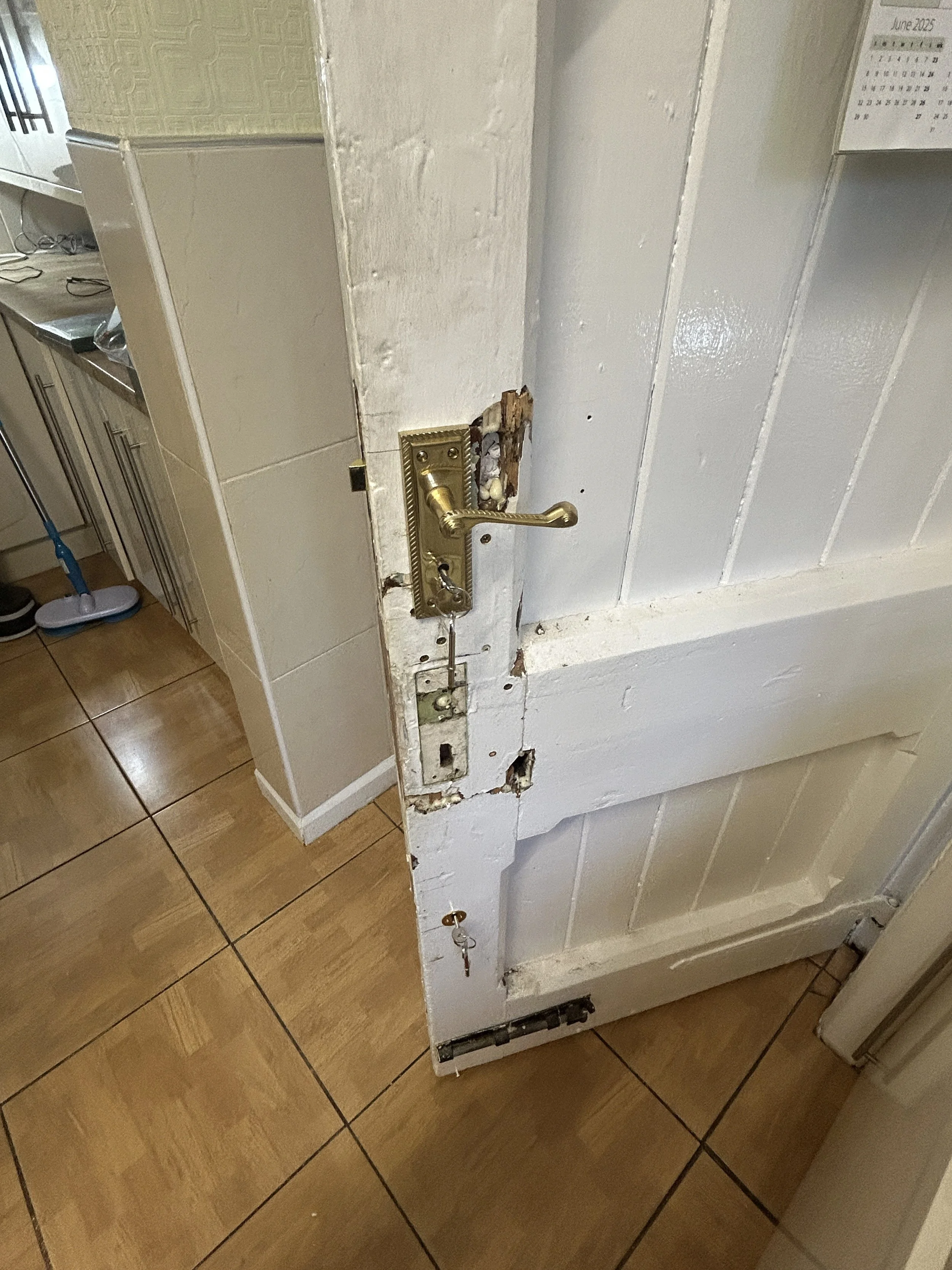 A partially removed door with a brass handle, revealing damaged doorframe and wall behind it, in a room with tiled floor and kitchen visible in the background.