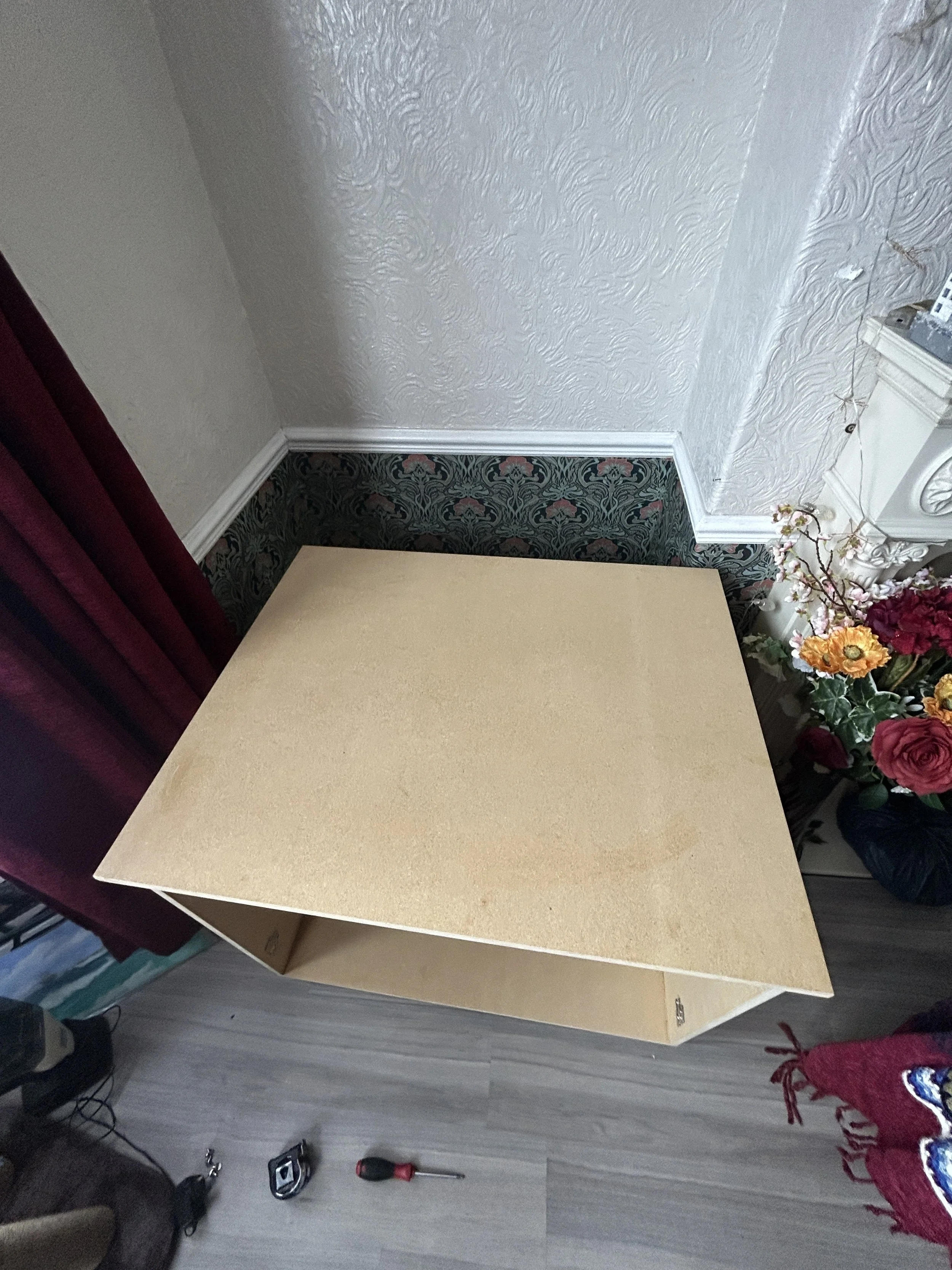 The image shows a piece of wood or particle board resting against a wall in a room with patterned wallpaper and floral decorations. There is a red curtain on the left side and some tools, including a screwdriver and a cordless drill, on the floor nea