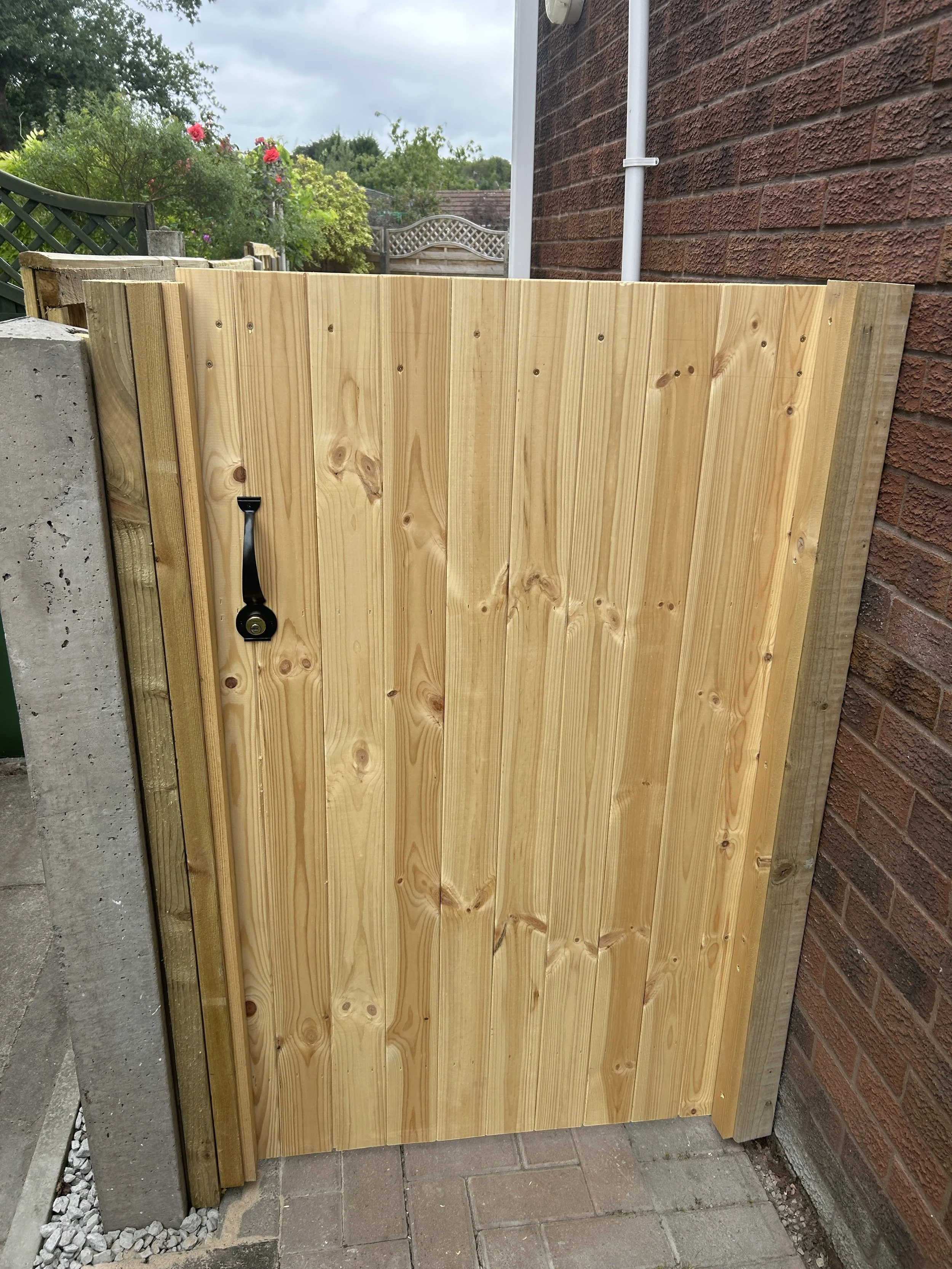 A newly installed wooden fence gate with a black latch, attached to a brick wall on the right, a concrete post on the left, and paving stones on the ground. There are gardening plants and a garden gate visible in the background under a cloudy sky.