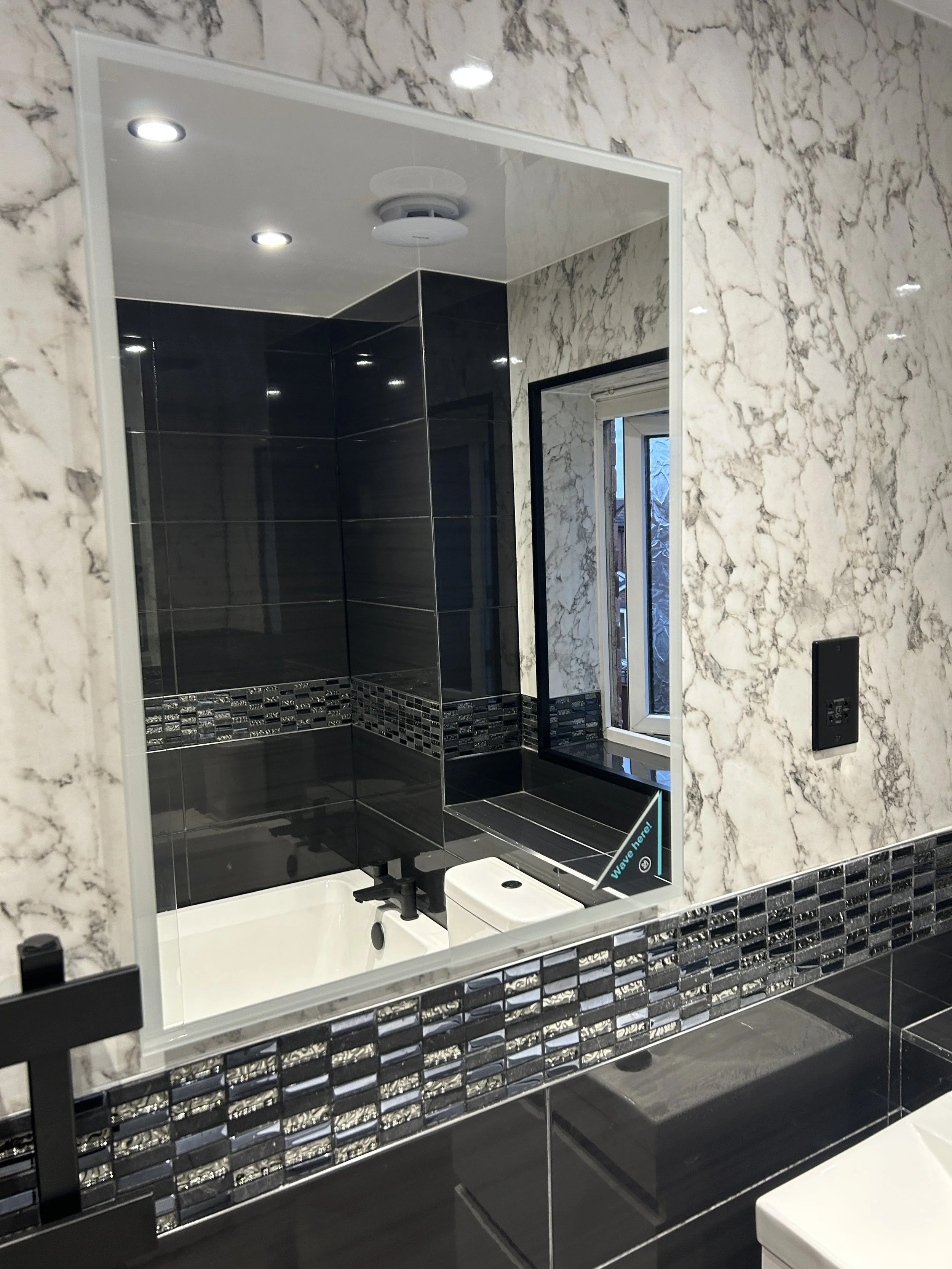 A bathroom mirror reflecting a corner with black and white tiled walls, a small window, and a sign that says 'Wave here!'