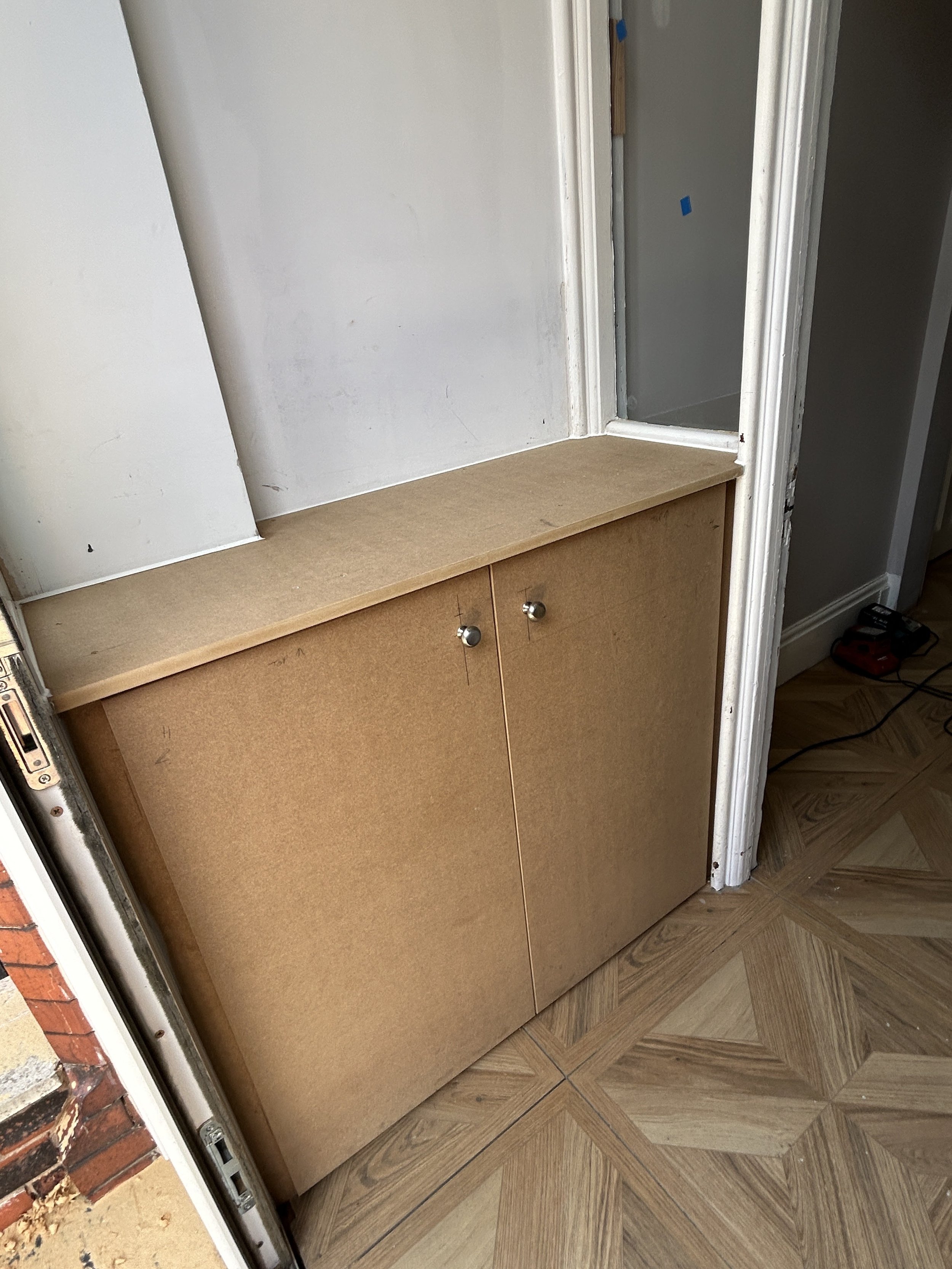 Wooden cabinet with two doors, placed against a wall with some paint or drywall repair tape visible near the doorway on the right. The floor has a wood-like patterned tile.